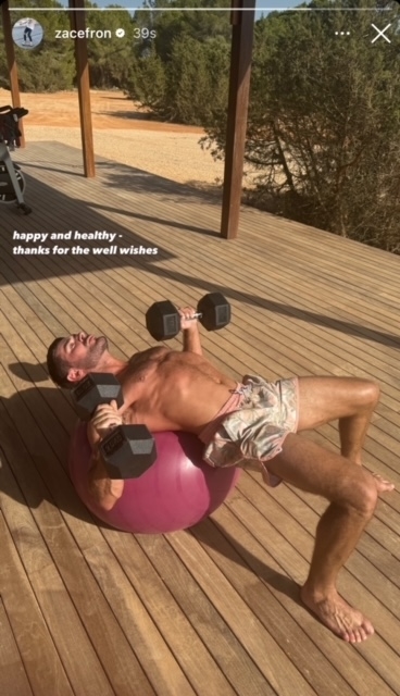 Zac Efron is outdoors, lying on an exercise ball while lifting dumbbells. He is shirtless, wearing shorts; text reads, "happy and healthy thanks for the well wishes."