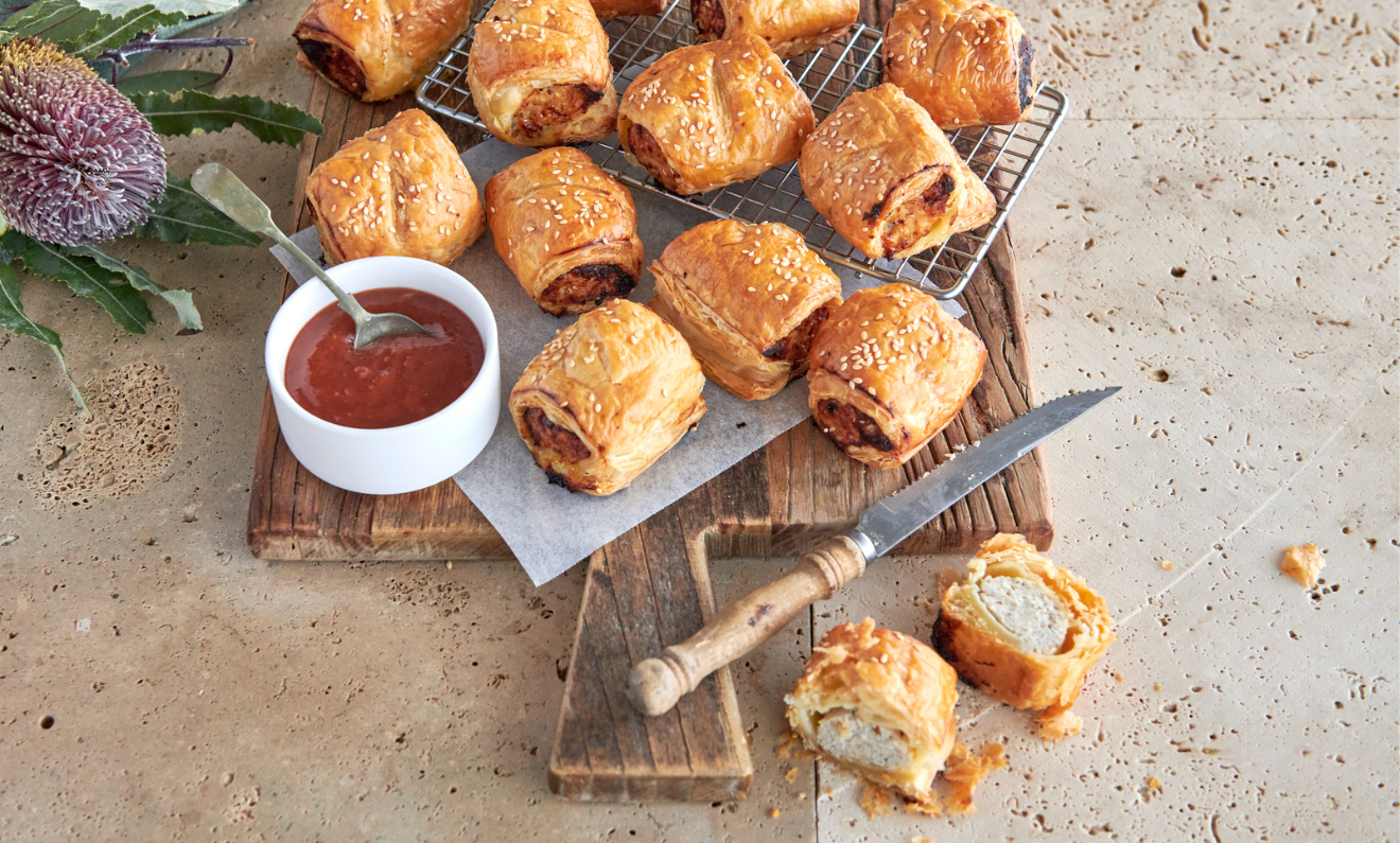 A wooden board with flaky sausage rolls, a white bowl of dipping sauce, a knife, and greenery on a textured surface