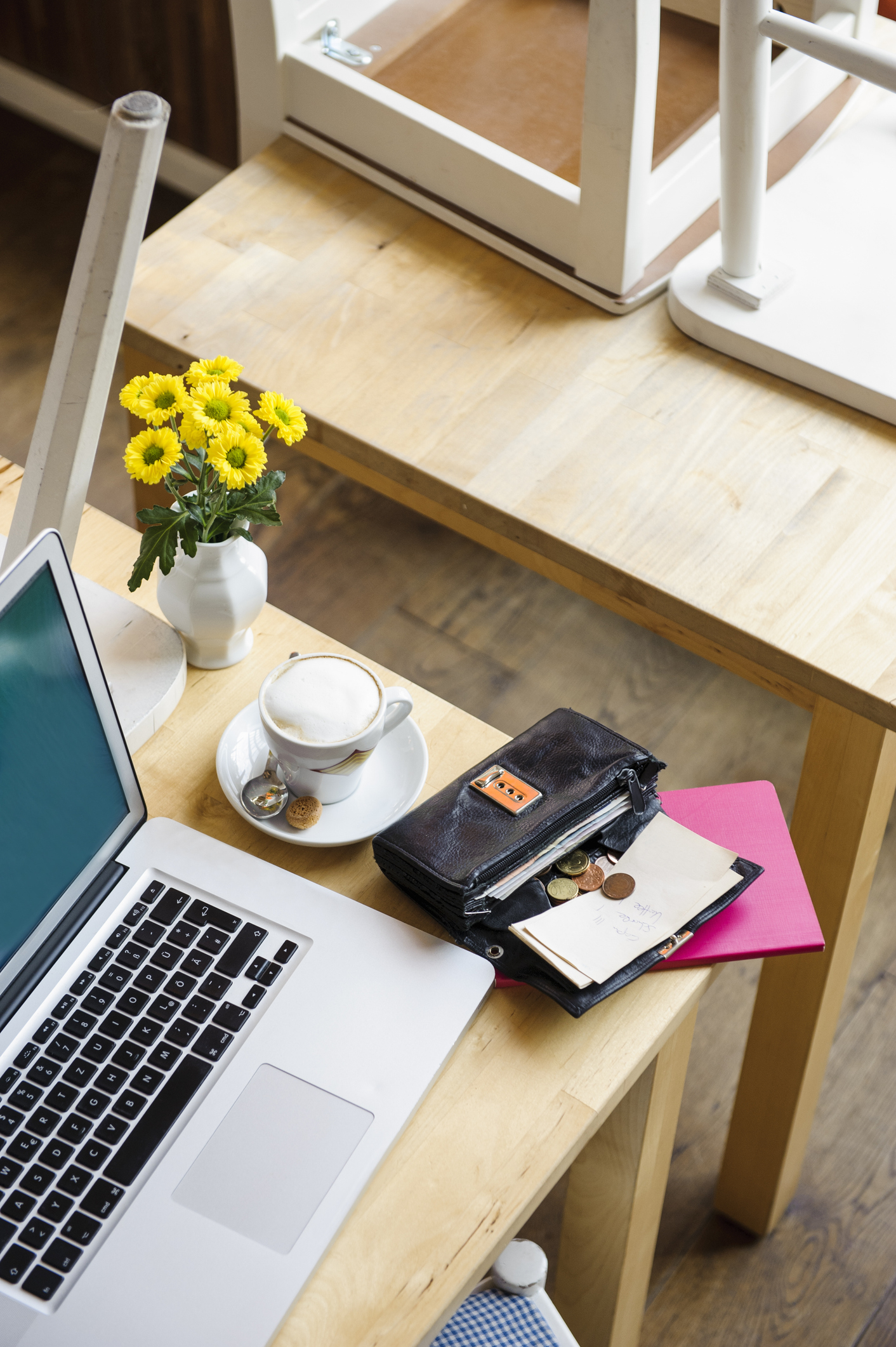 A laptop, a wallet, coins, a notebook, and a vase with yellow flowers on a wooden table with a cup of coffee