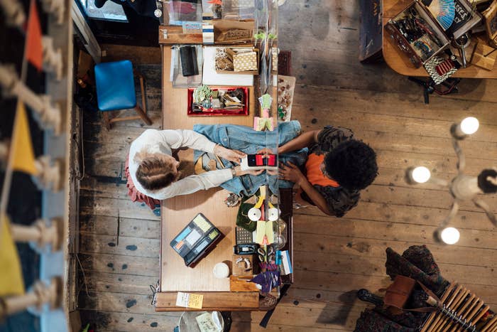 Two people, one with light-colored hair and one with dark hair, are seated at a cluttered table exchanging items in a room filled with various objects