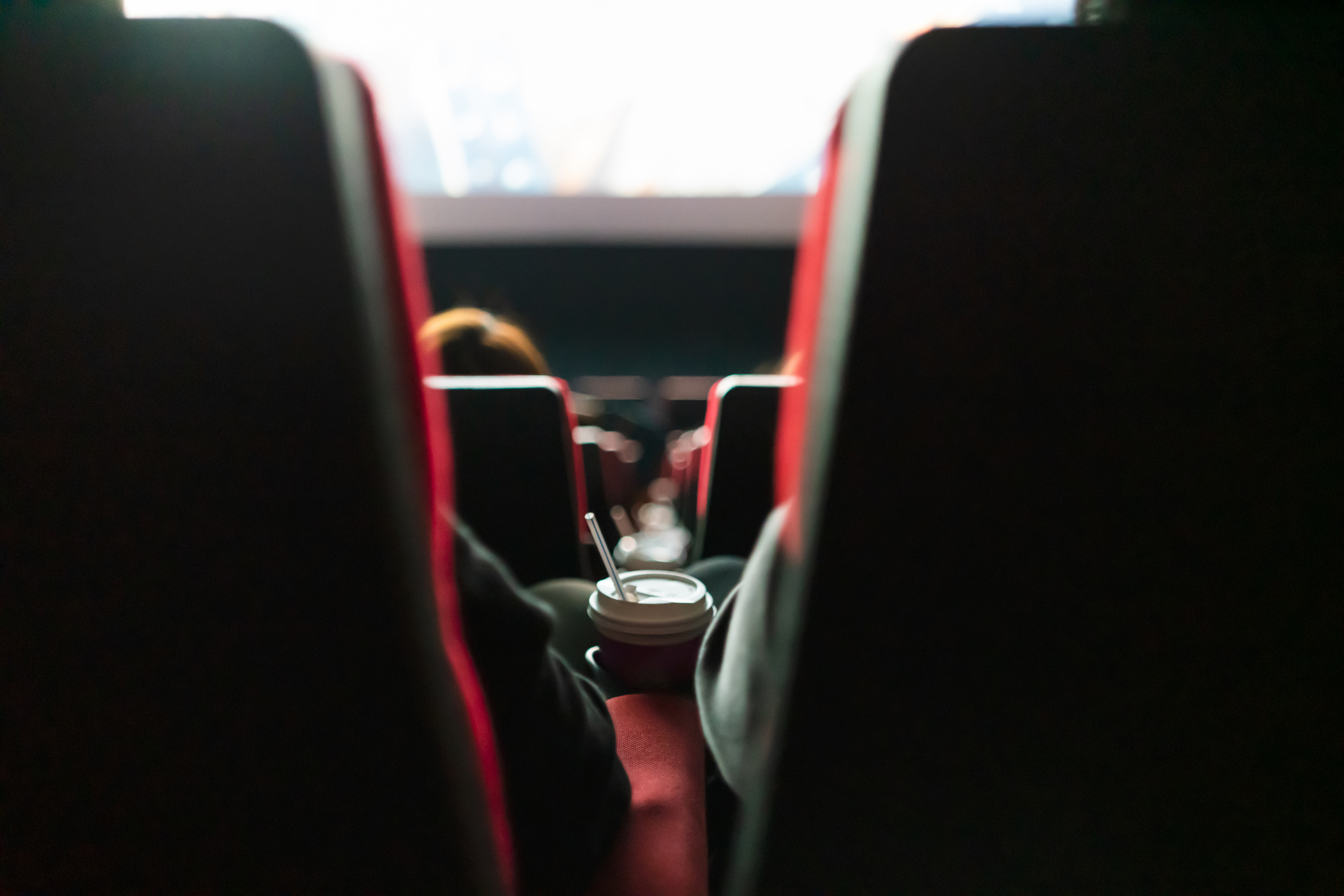People seated in a movie theater, viewed from behind, with one person holding a cup with a straw on the armrest. The movie screen is visible in the background