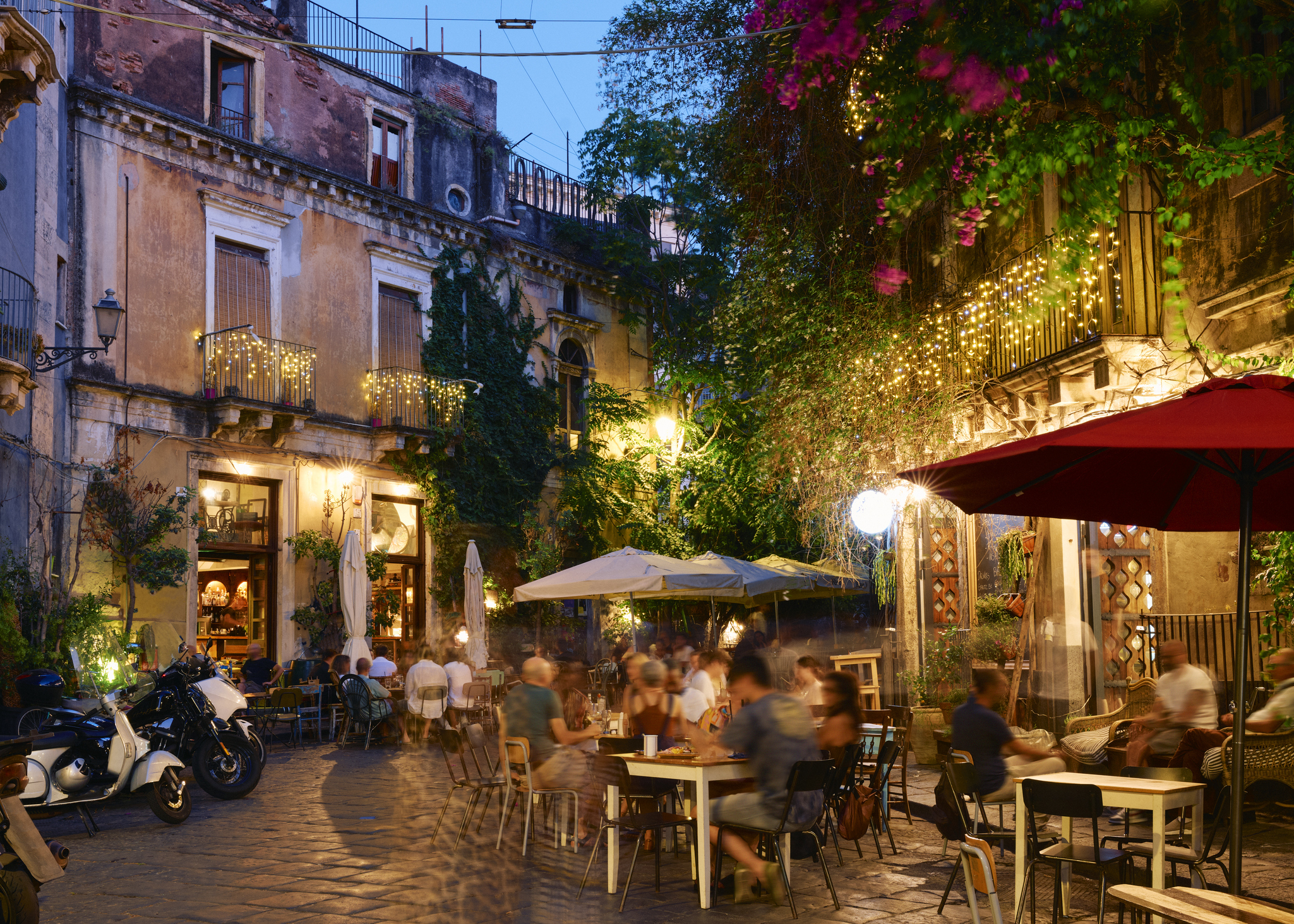 People dining at an outdoor cafe in a picturesque European street with buildings covered in greenery and lights. Some tables are under umbrellas