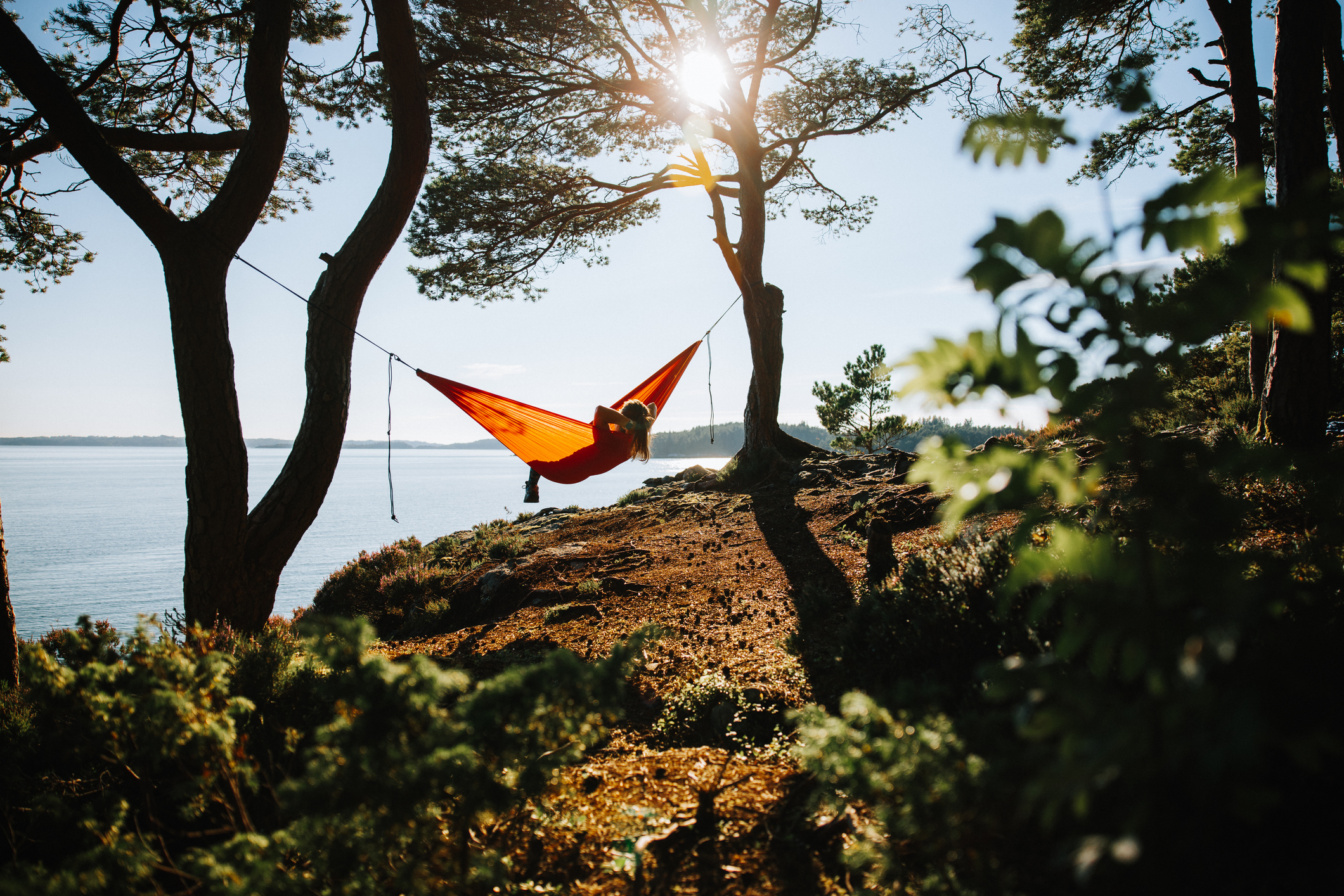 Person relaxing in a hammock tied between trees on a rocky shoreline with a calm sea in the background. Sunlight filters through the treetops