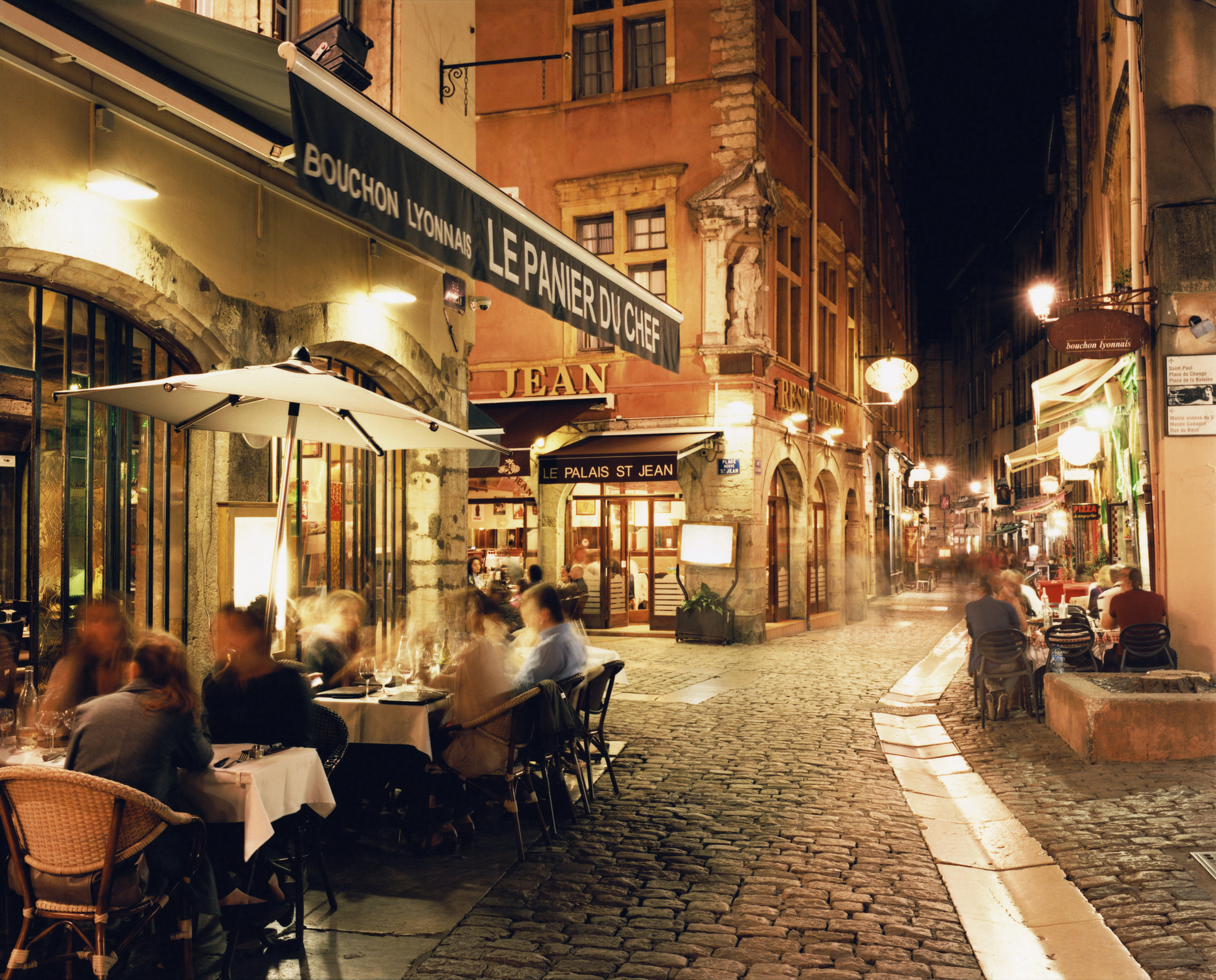 People dining outdoors on a cobblestone street at a French café named "Le Panier du Chef" at night, with warm ambient lighting enhancing the historic architecture
