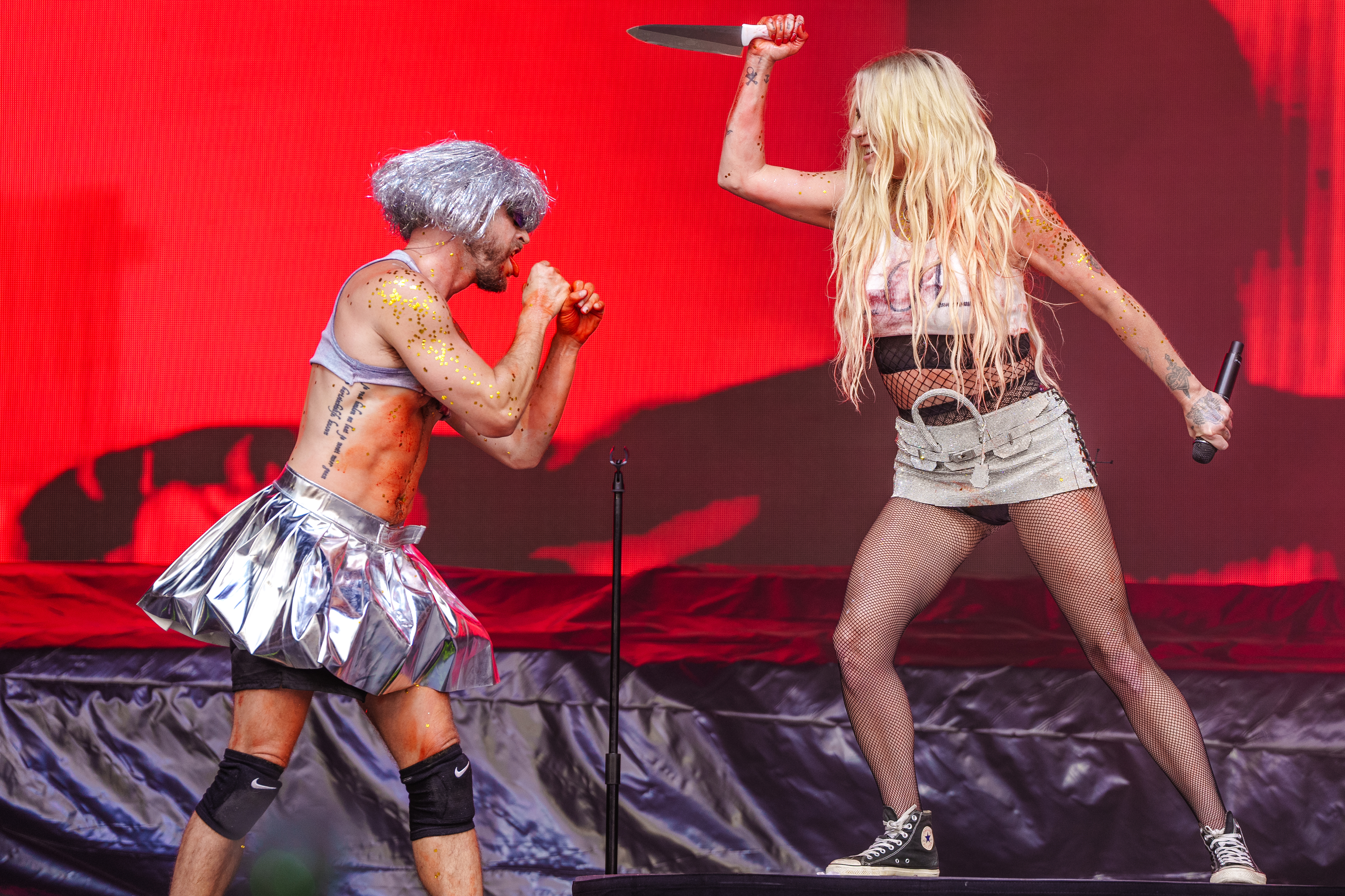 Ke$ha performs on stage with a male dancer dressed in a metallic skirt. She holds a prop knife above her head