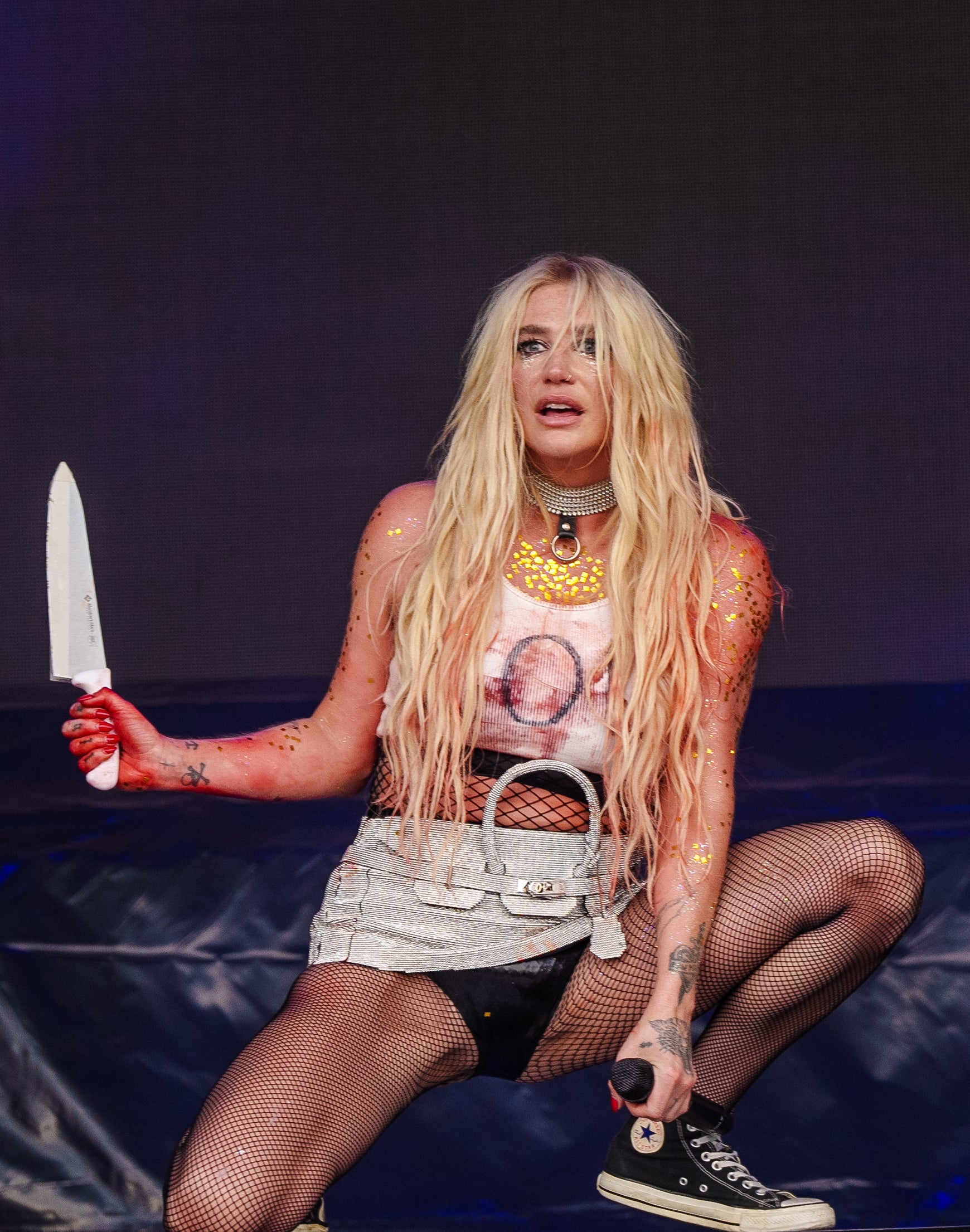 Kesha performs on stage, crouching with a knife. She wears a graphic tank top, metallic skirt, fishnet stockings, and Converse shoes