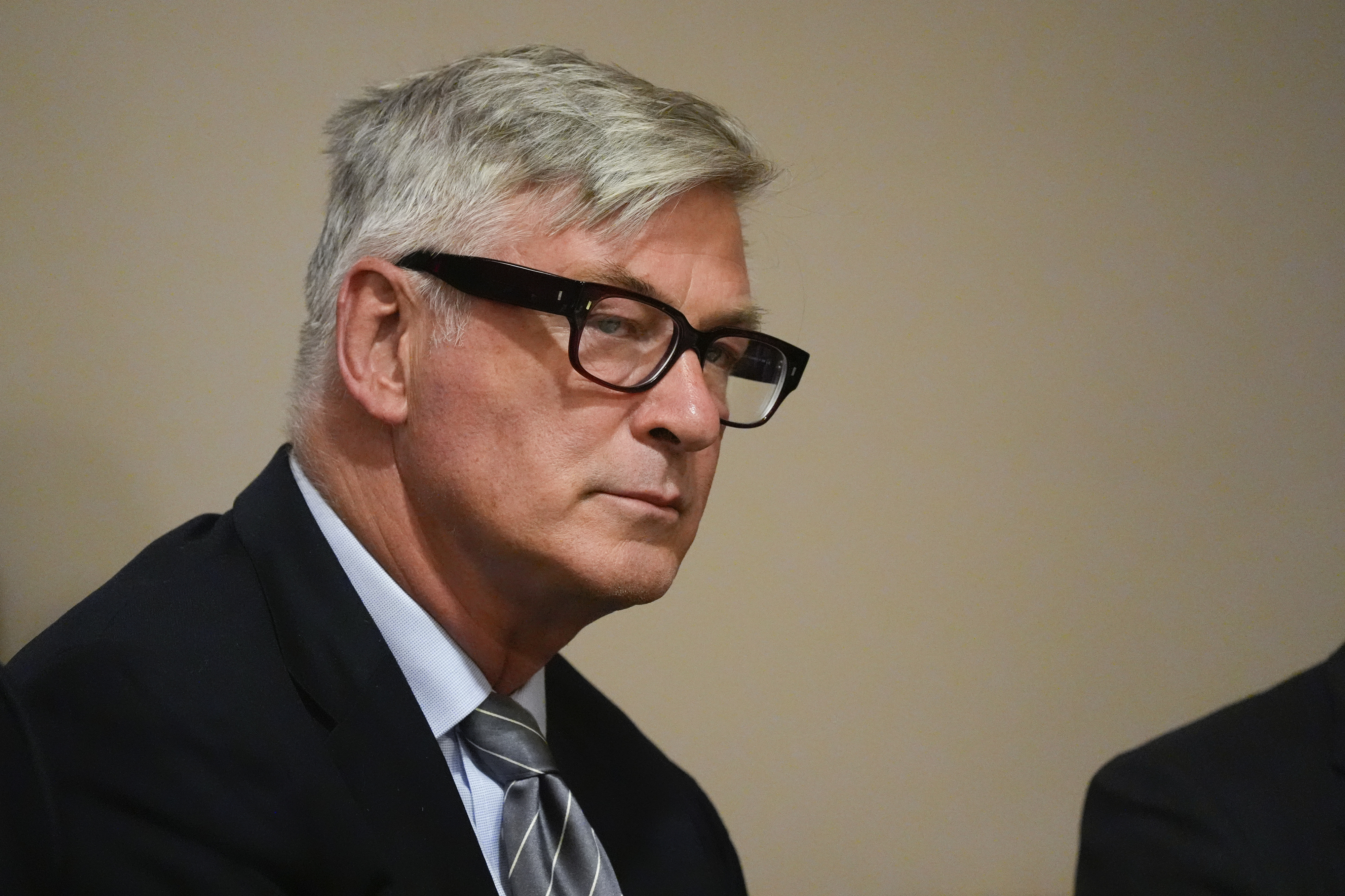 Alec Baldwin is wearing glasses, a dark suit, and a striped tie while looking to the side