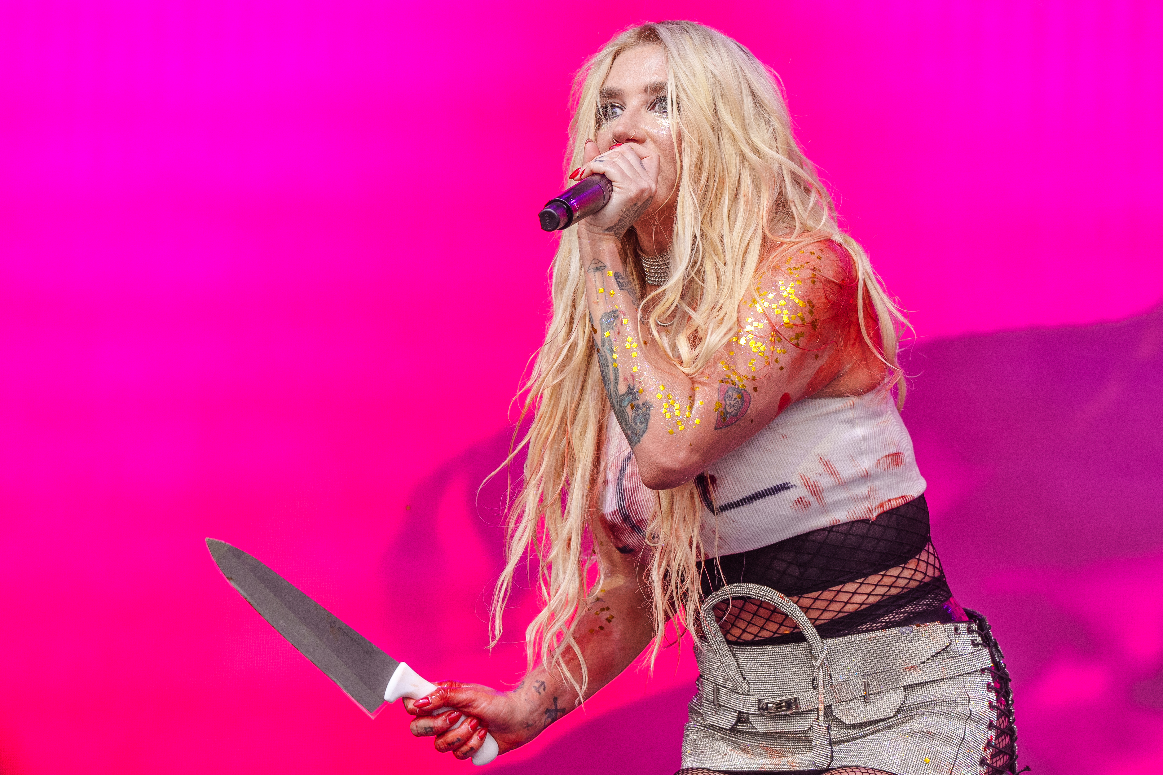 Kesha performs on stage holding a knife, wearing a distressed outfit with rips and glitter paint accents