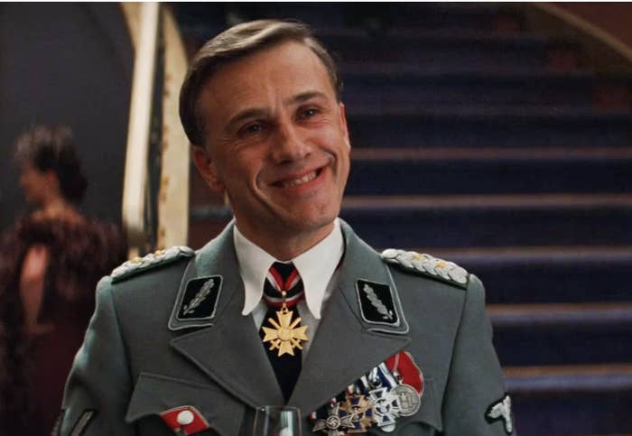 Christoph Waltz in military uniform, smiling while holding a glass, on a staircase with another person blurred in the background