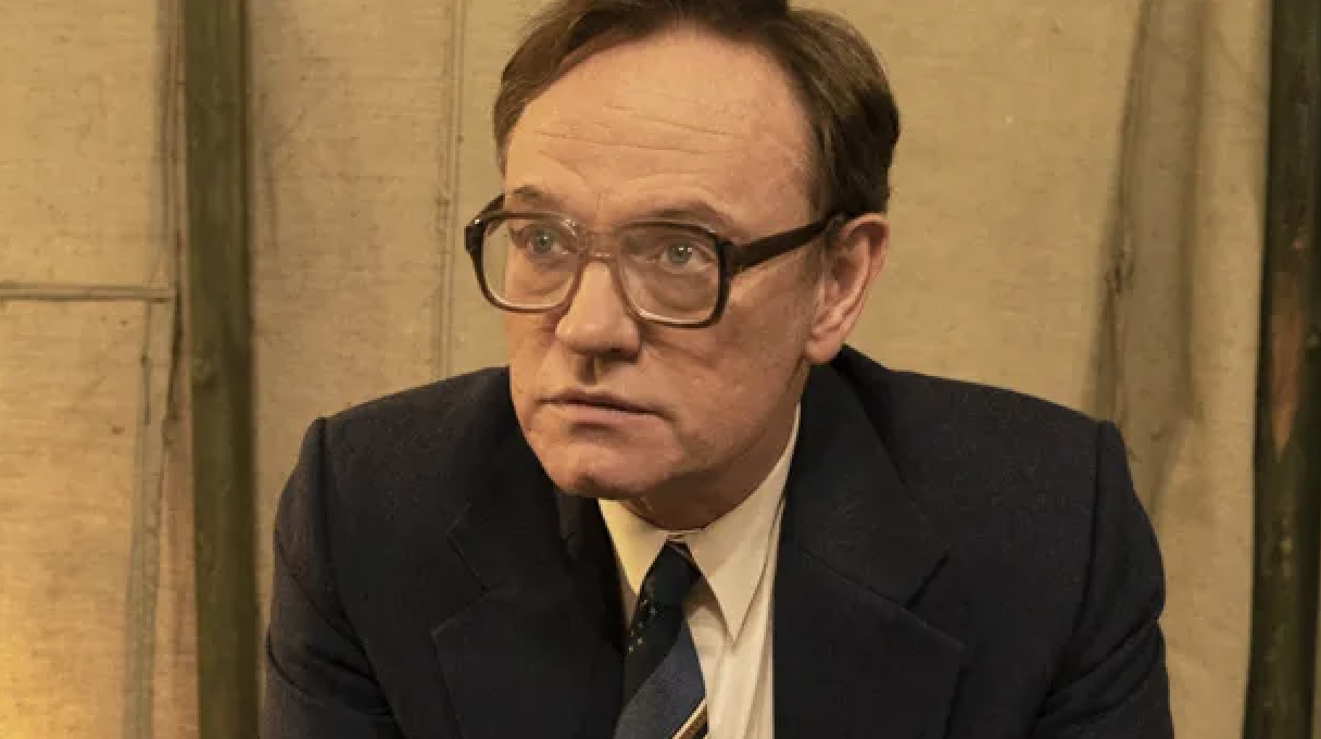 A man is sitting and looking slightly off to the side. He is wearing a suit, tie, and glasses