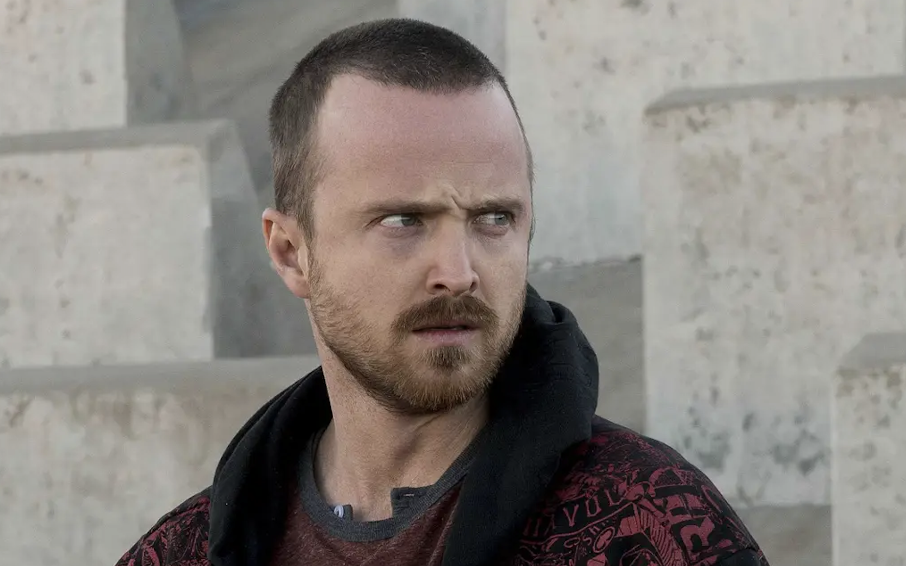 Aaron Paul looking to the side with a serious expression, wearing a dark hooded jacket with a patterned design and a T-shirt underneath