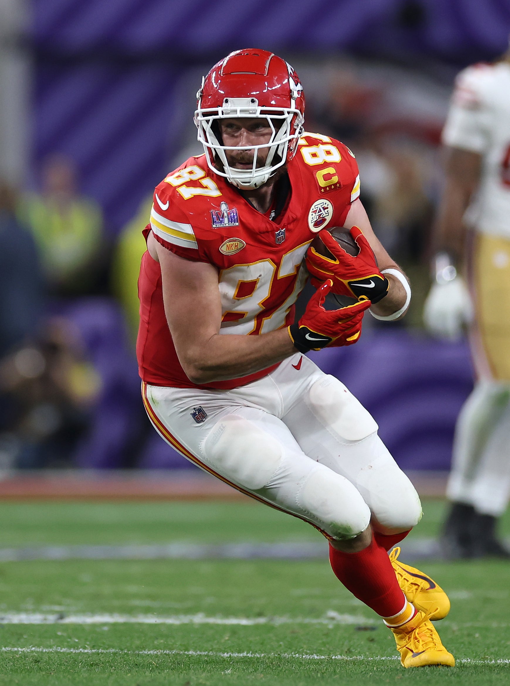 Travis Kelce, in football gear, runs with the ball during a game. Opposing team members are visible in the background