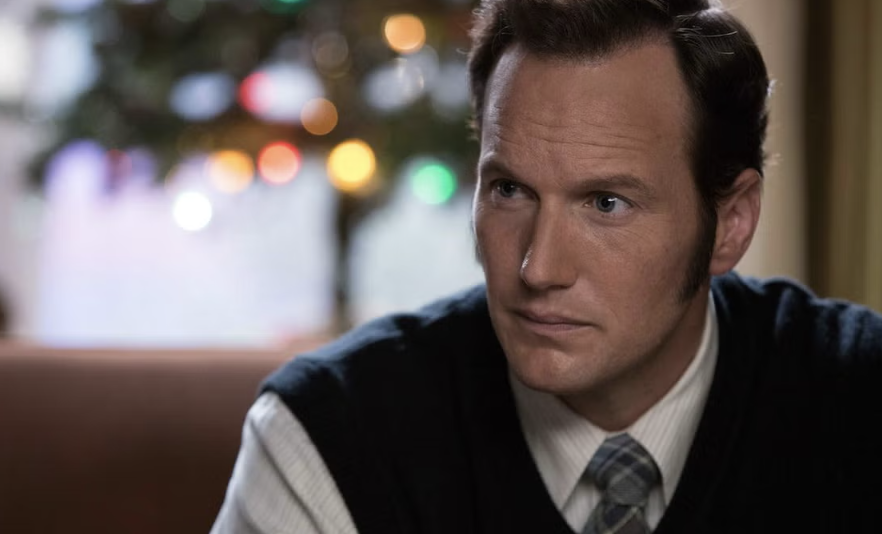 Patrick Wilson wearing a collared shirt and a tie, sitting indoors with blurred lights in the background