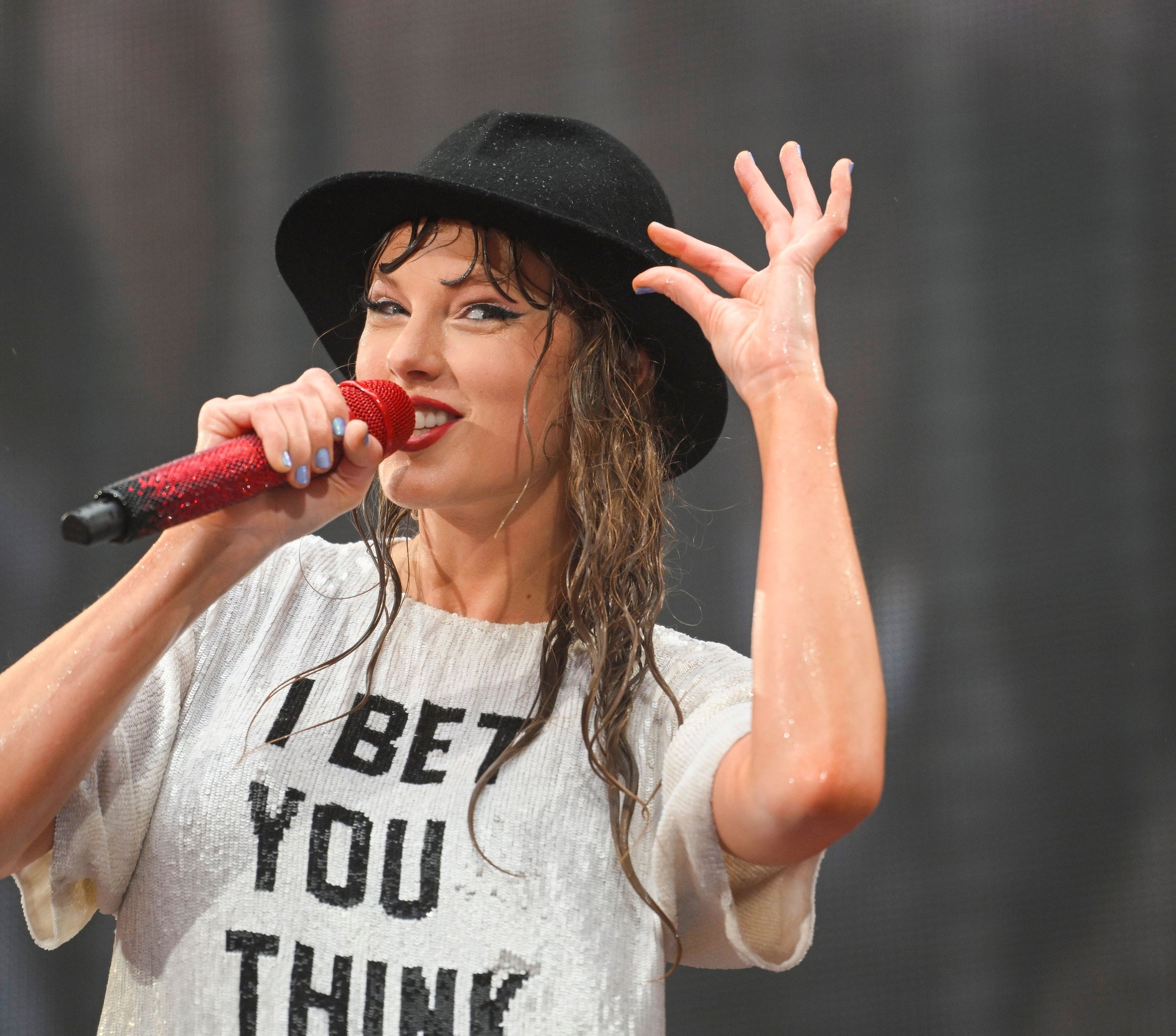 Taylor Swift, with wet hair under a black hat, sings into a red microphone and wears a white shirt that reads &quot;I BET YOU THINK ABOUT ME.&quot;