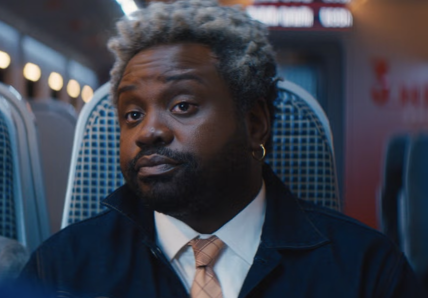 Brian Tyree Henry sits on a train, wearing a tie, white dress shirt, and dark jacket, with a neutral expression