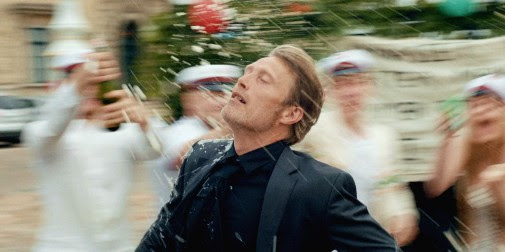 Mads Mikkelsen, in a suit, gets splashed by liquid during a celebration with people in the background