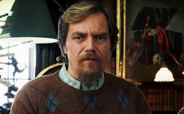 Michael Shannon in an argyle sweater, seated indoors with a focused expression. A lamp and art piece are in the background