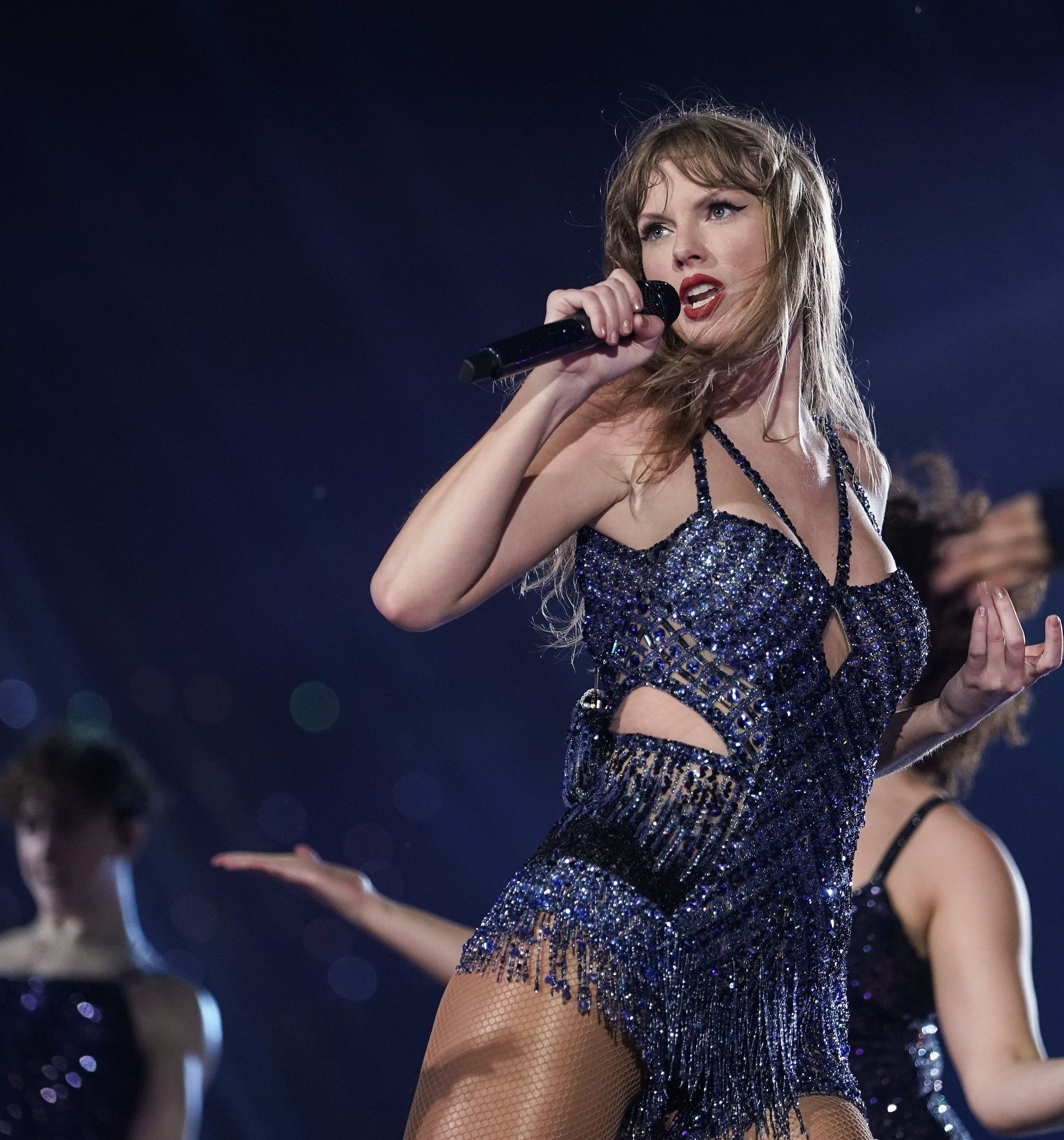 Taylor Swift performing on stage in a sparkly, beaded outfit with backup dancers in the background. She is holding a microphone and gesturing dynamically