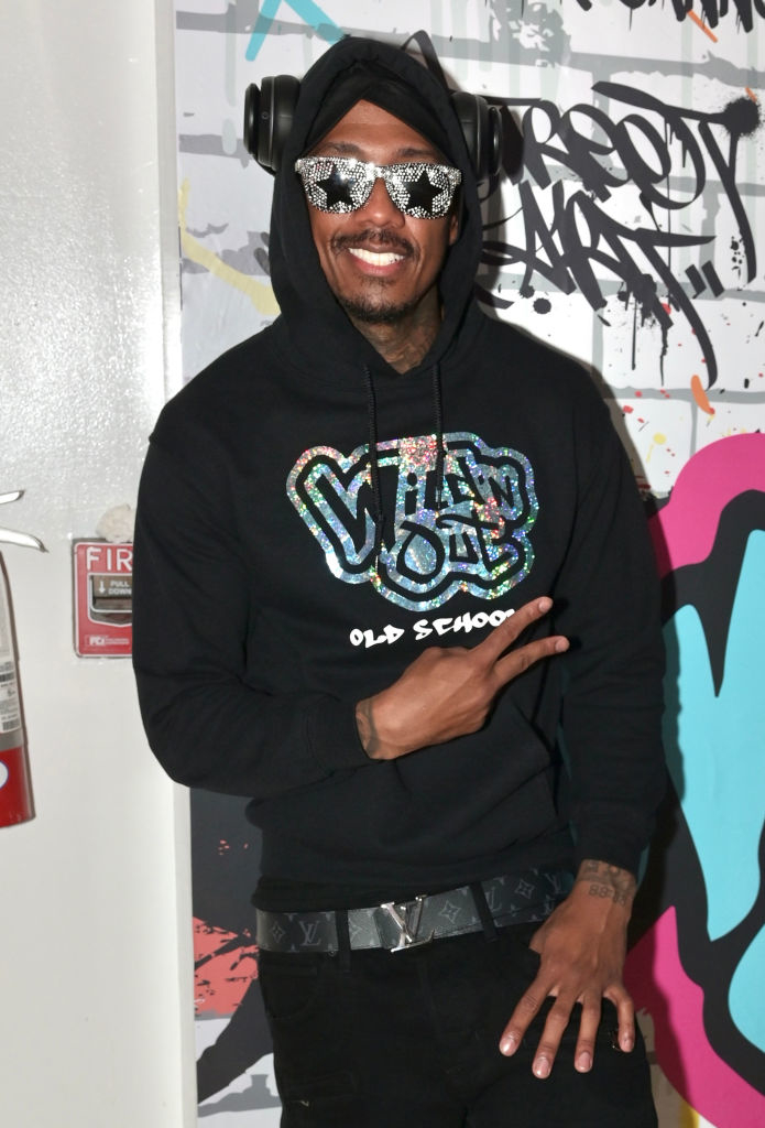 Nick Cannon wearing a Wild 'N Out hoodie, futuristic sunglasses, headphones, and a belt with a metallic buckle, posing with a peace sign. Graffiti backdrop in the background