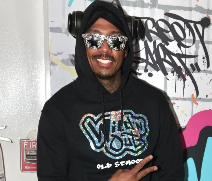 Nick Cannon wearing a Wild 'N Out hoodie, futuristic sunglasses, headphones, and a belt with a metallic buckle, posing with a peace sign. Graffiti backdrop in the background