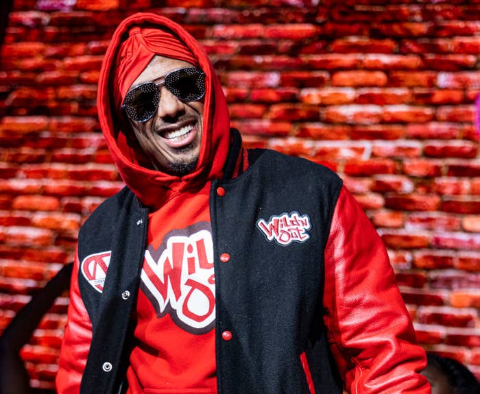 Nick Cannon wearing a stylish "Wild 'N Out" varsity jacket and hoodie, with sunglasses, smiling while standing in front of a stage background