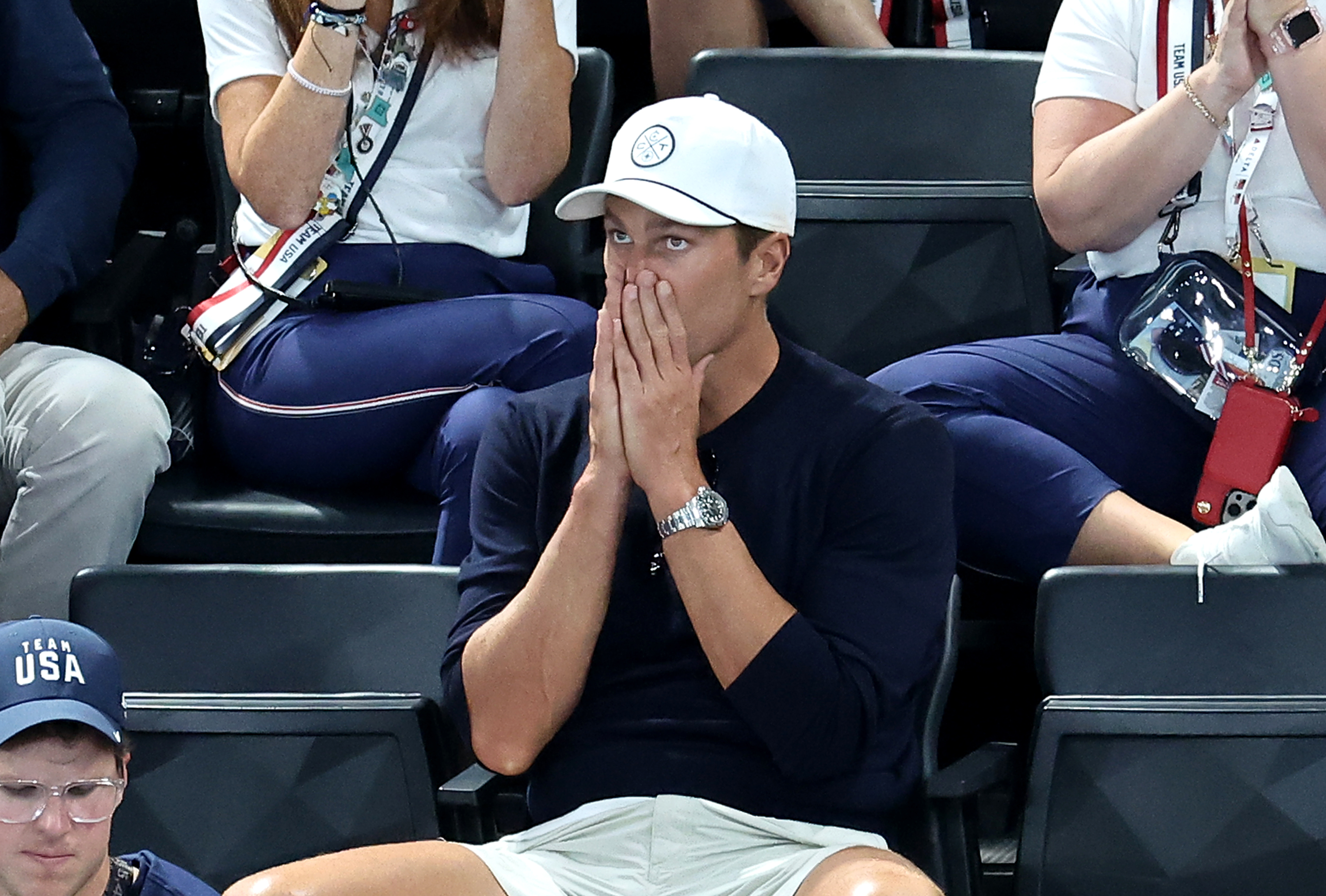 Unidentified person wearing a white cap and dark blue shirt sits in the audience with hands over mouth, looking distressed. Others around them are clapping or watching