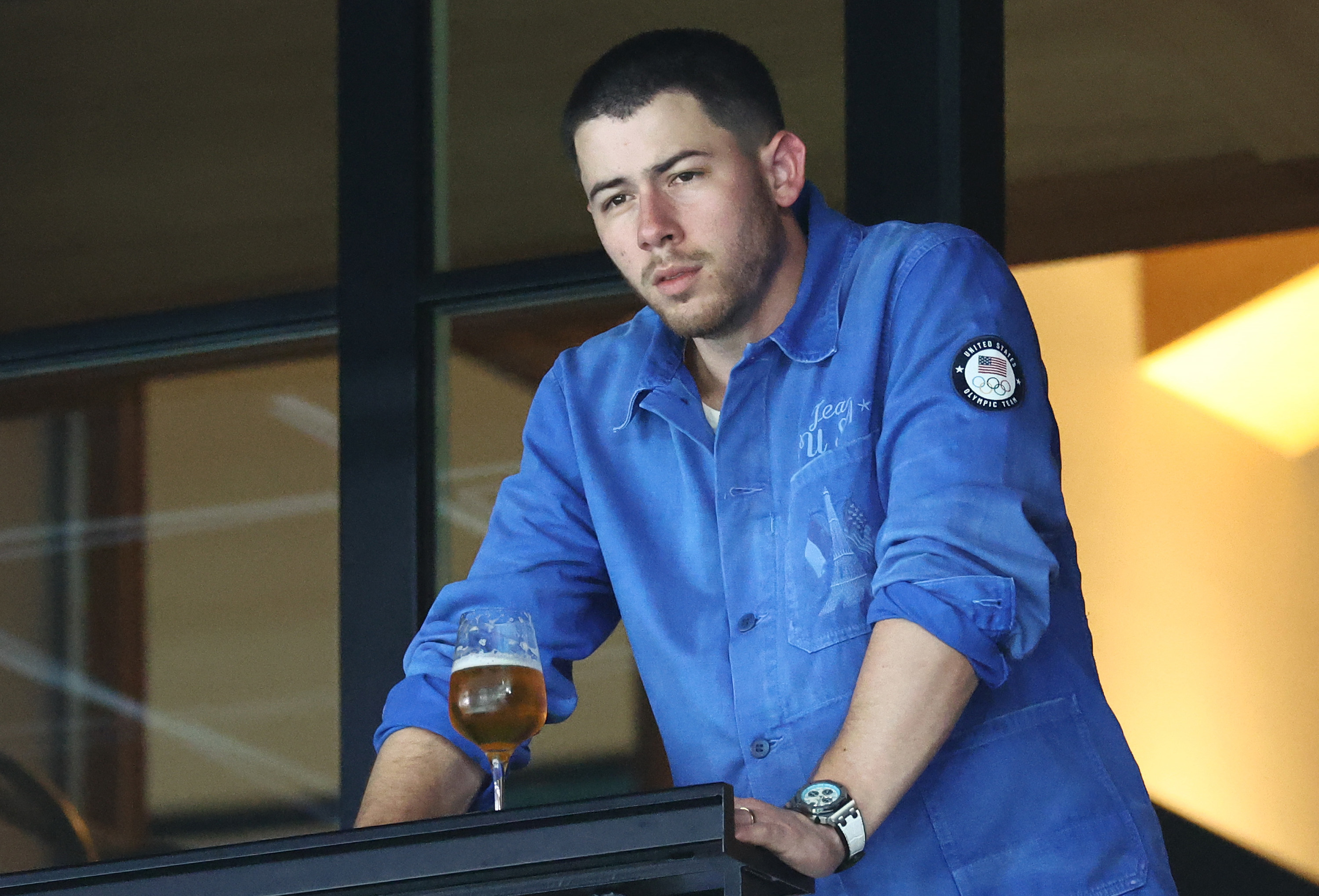 Nick Jonas in a casual, blue button-down shirt, holding a drink and looking thoughtful