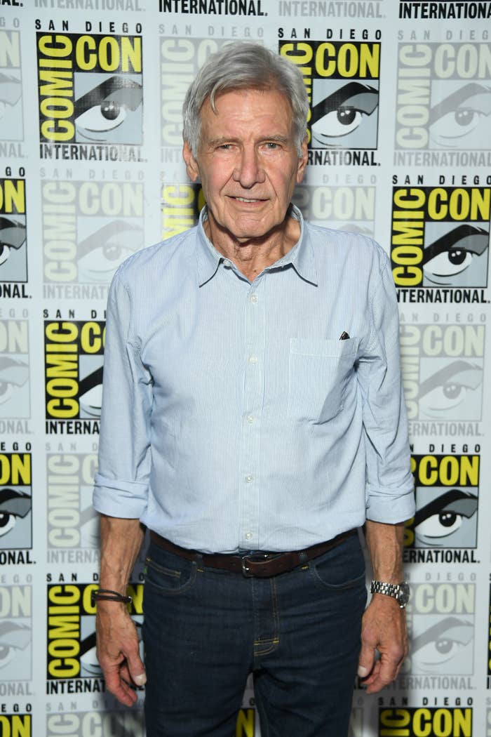 Harrison Ford at San Diego Comic-Con International, standing in front of the event's branded backdrop. He is wearing a casual button-up shirt and jeans
