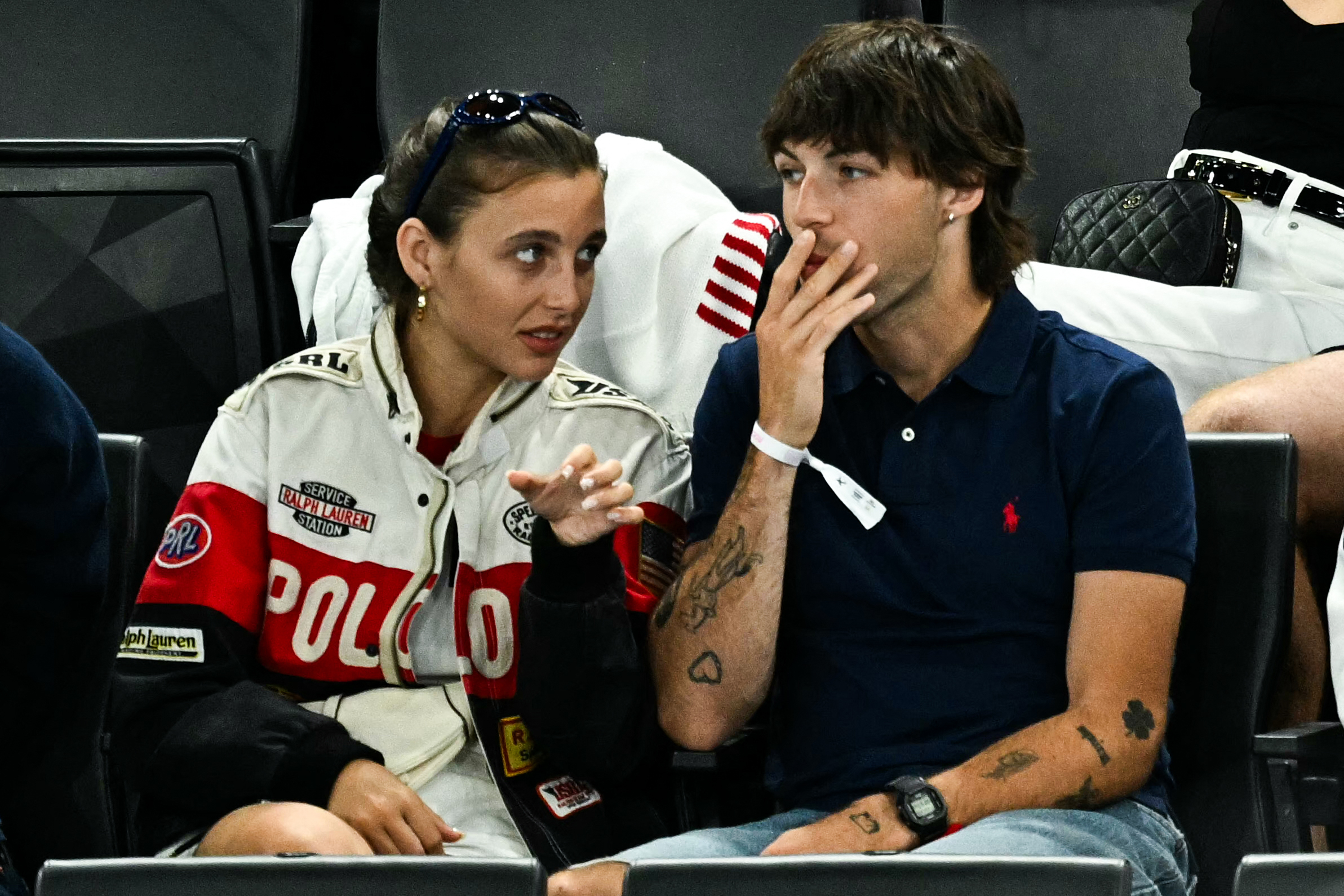 Emma Chamberlain and Role Model (Tucker Pillsbury) sit together at a sports event, engaged in conversation. Emma wears a racing jacket while Role Model wears a navy polo