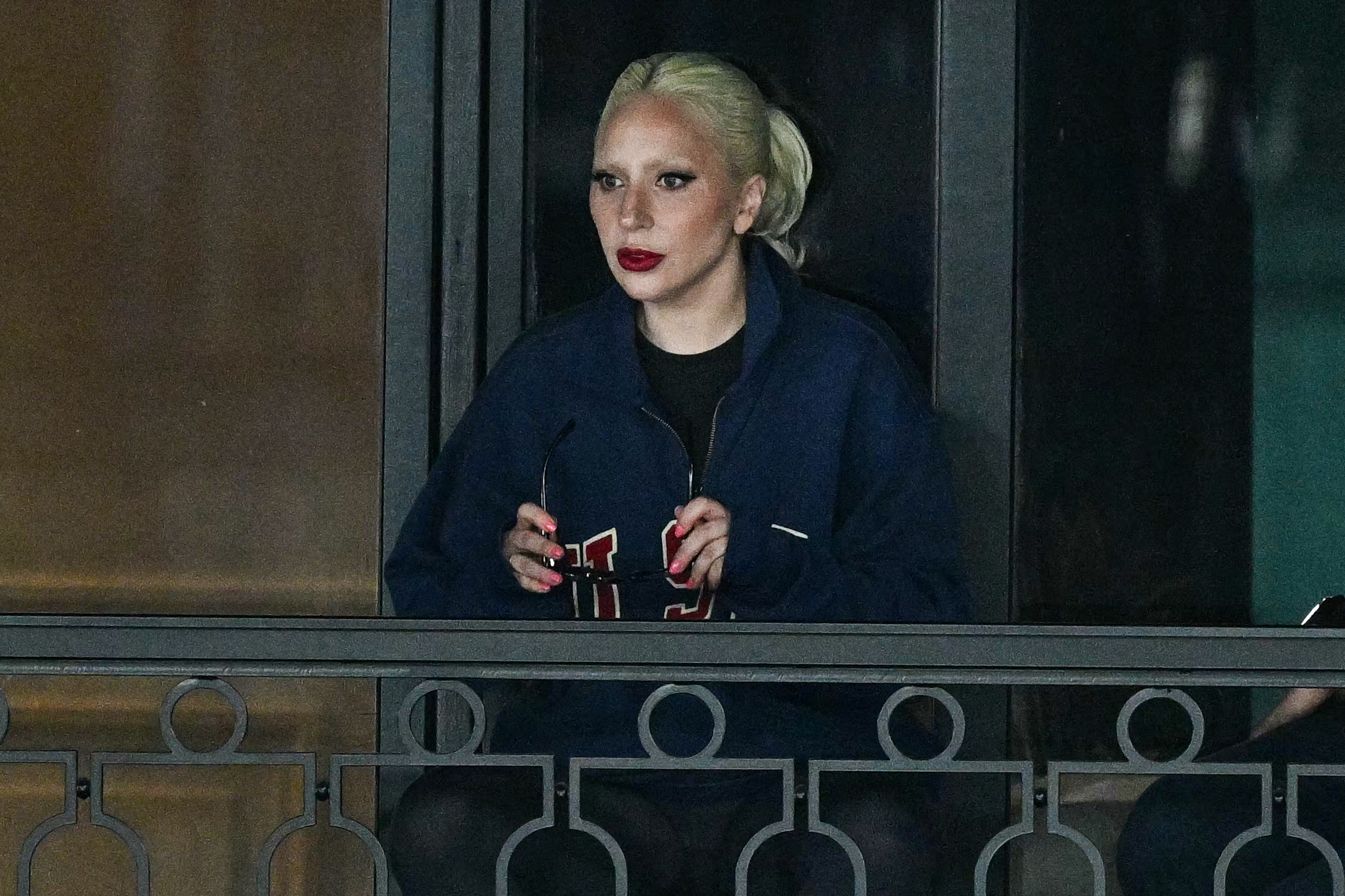 Lady Gaga holding sunglasses and dressed in casual attire, standing on a balcony