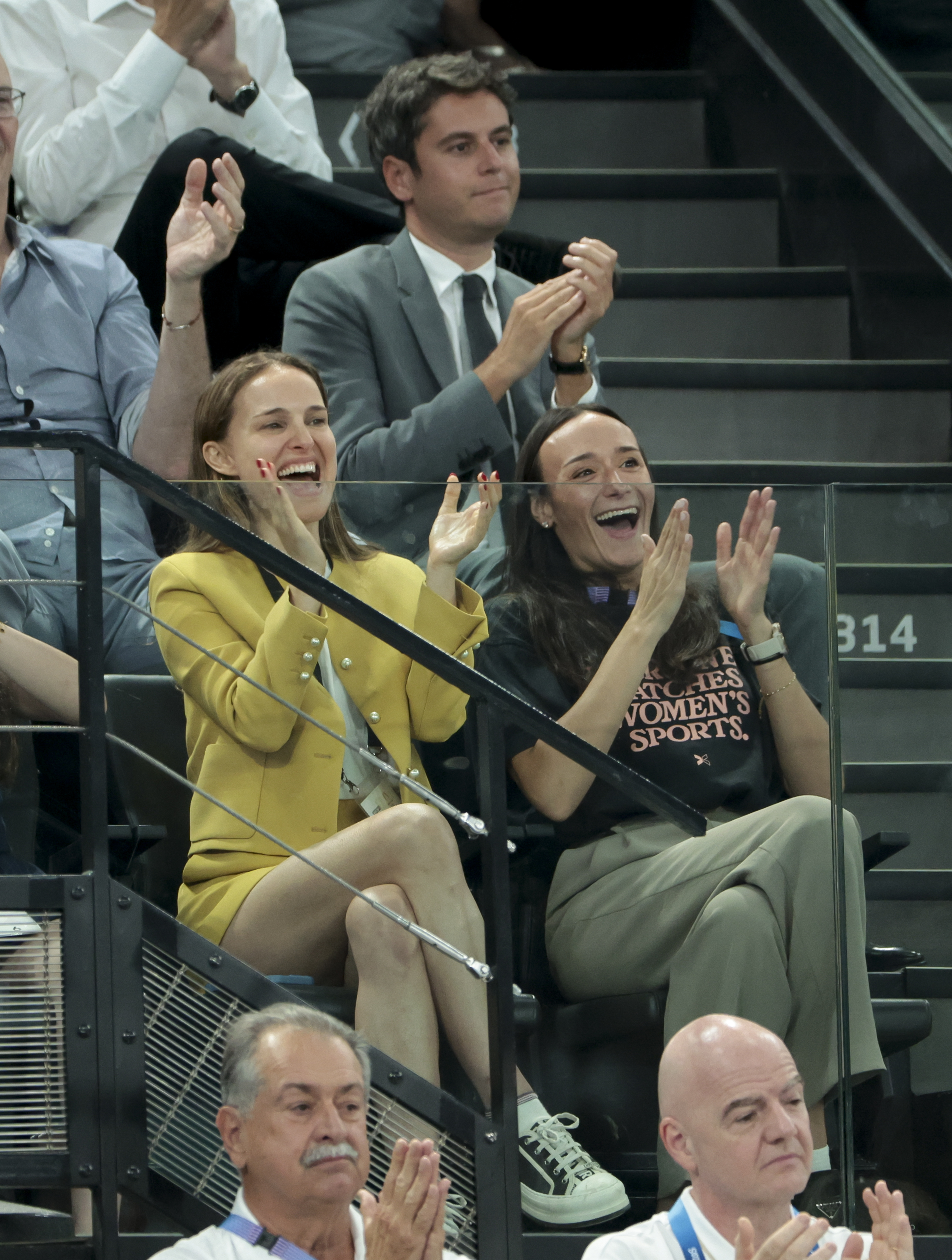 Natalie Portman and Kelley O'Hara cheer enthusiastically from the stands at a sports event, surrounded by other spectators