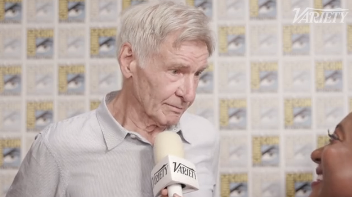Harrison Ford is being interviewed at a Variety event. He is wearing a light-colored, casual button-up shirt