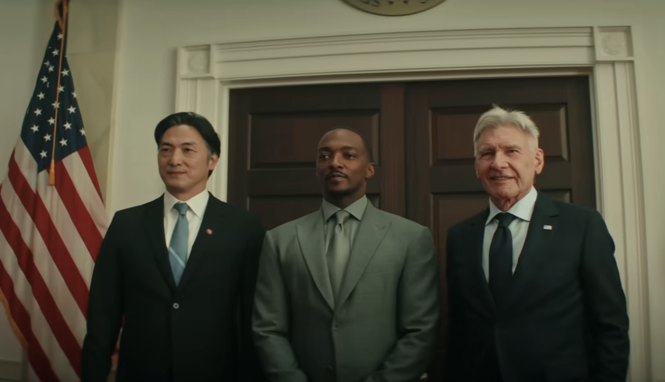 Ken Takemoto, Anthony Mackie, and Harrison Ford standing together in formal attire in front of a closed door and the American flag