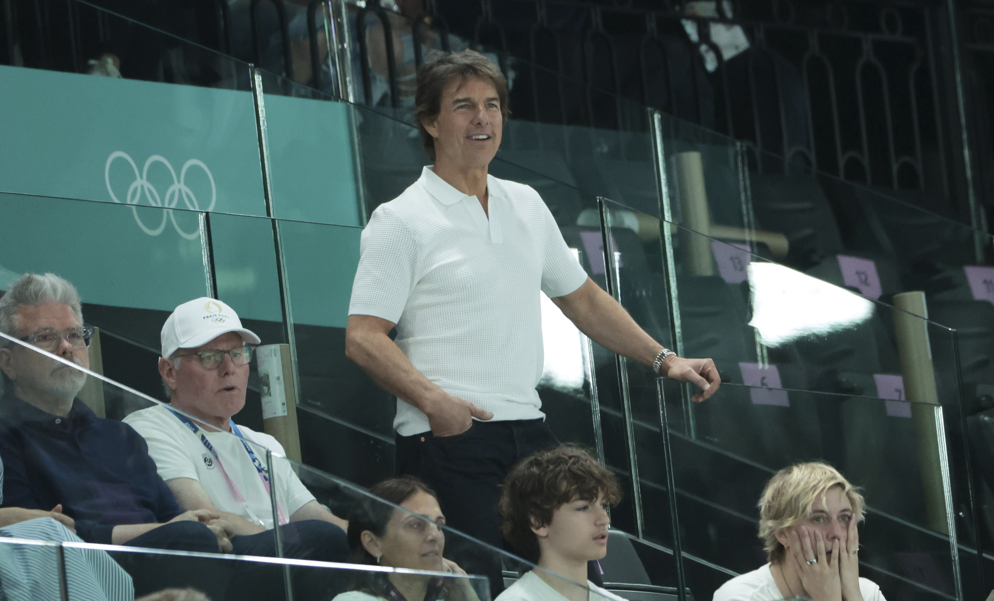 Tom Cruise stands and watches a sports event surrounded by other spectators. Olympic rings can be seen in the background