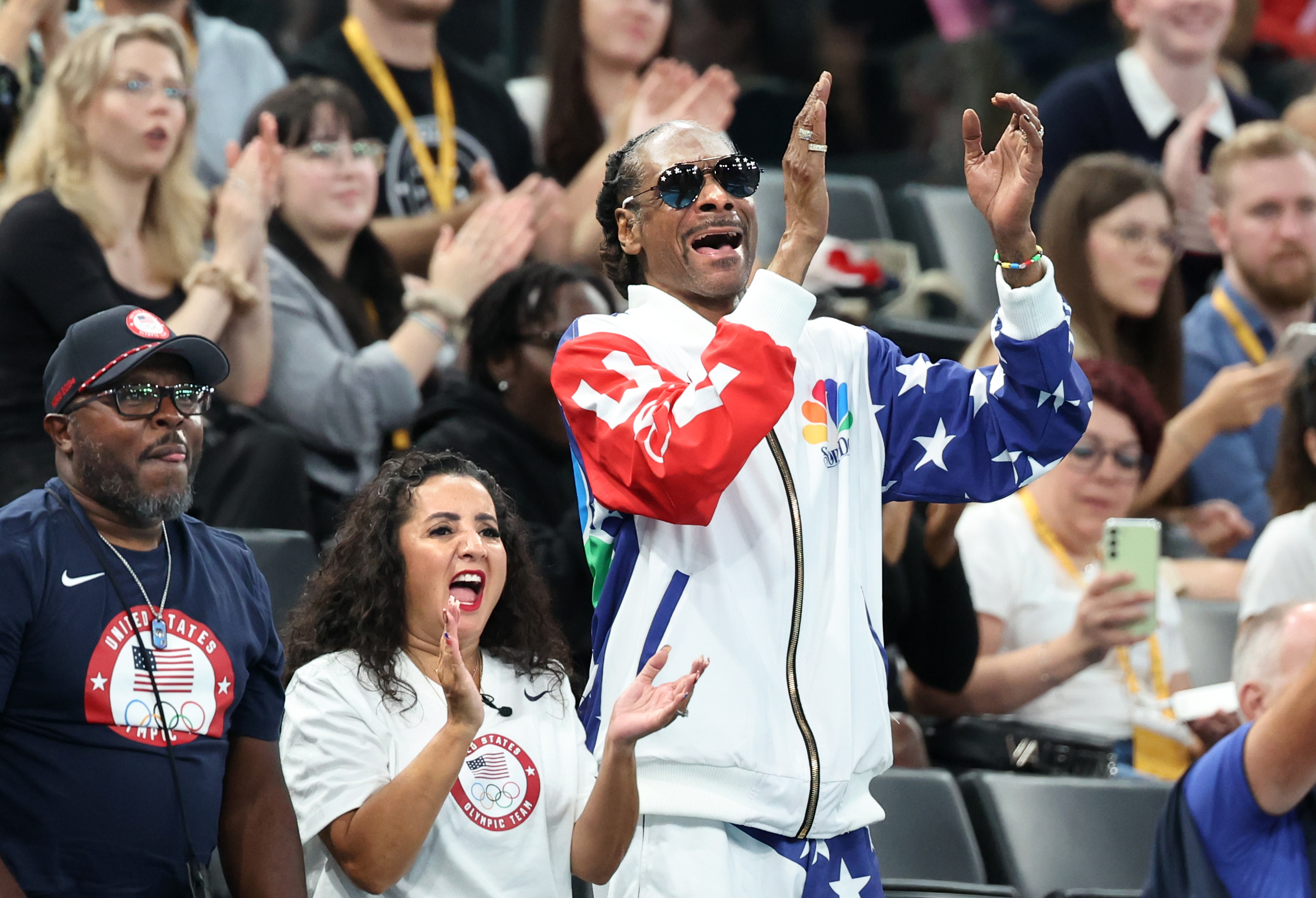 Snoop Dogg claps enthusiastically in the stands at an event, wearing a star-patterned tracksuit and sunglasses. Crowd members around him also cheer