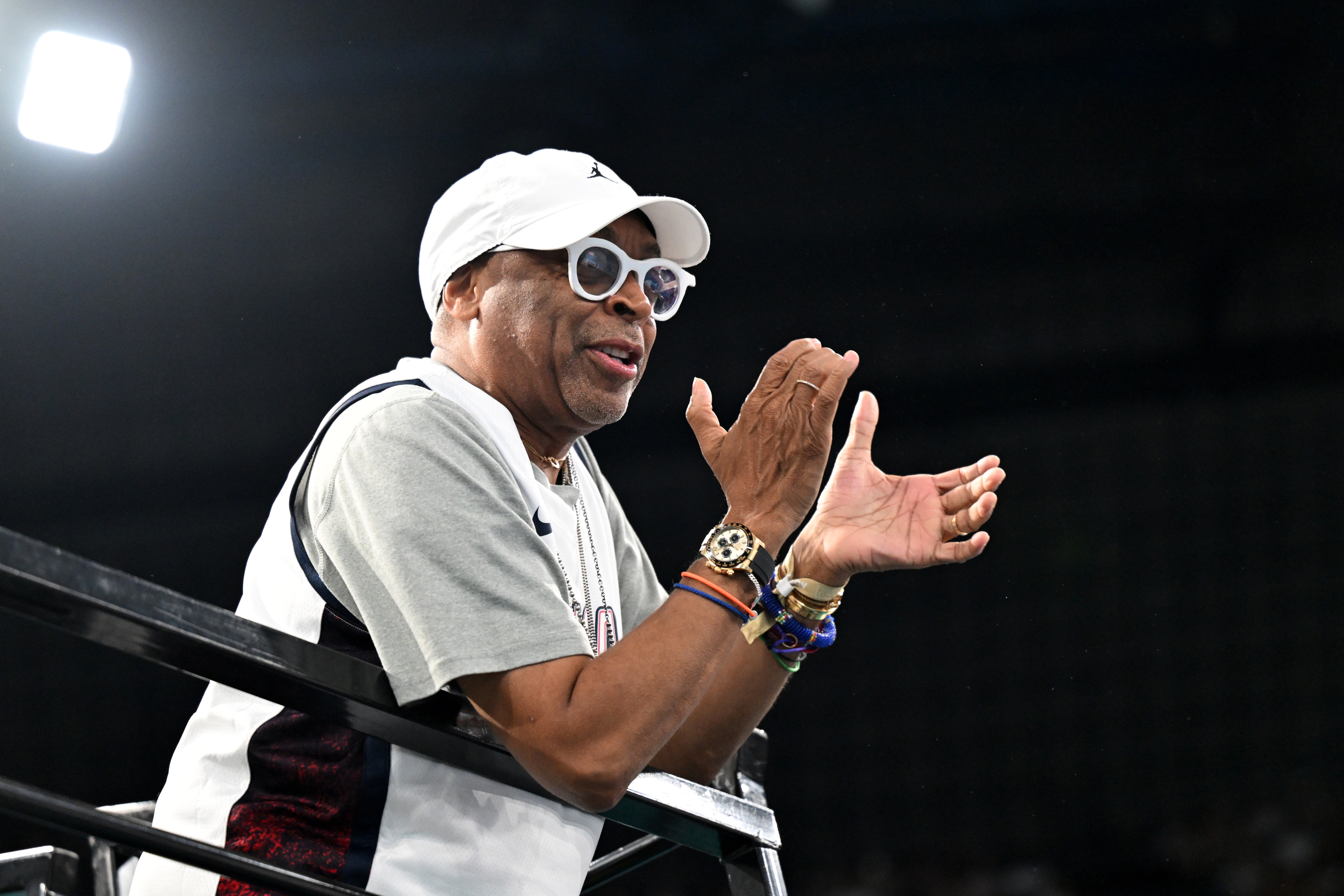 Spike Lee clapping at a sports event, wearing a white cap, round sunglasses, and casual athletic wear