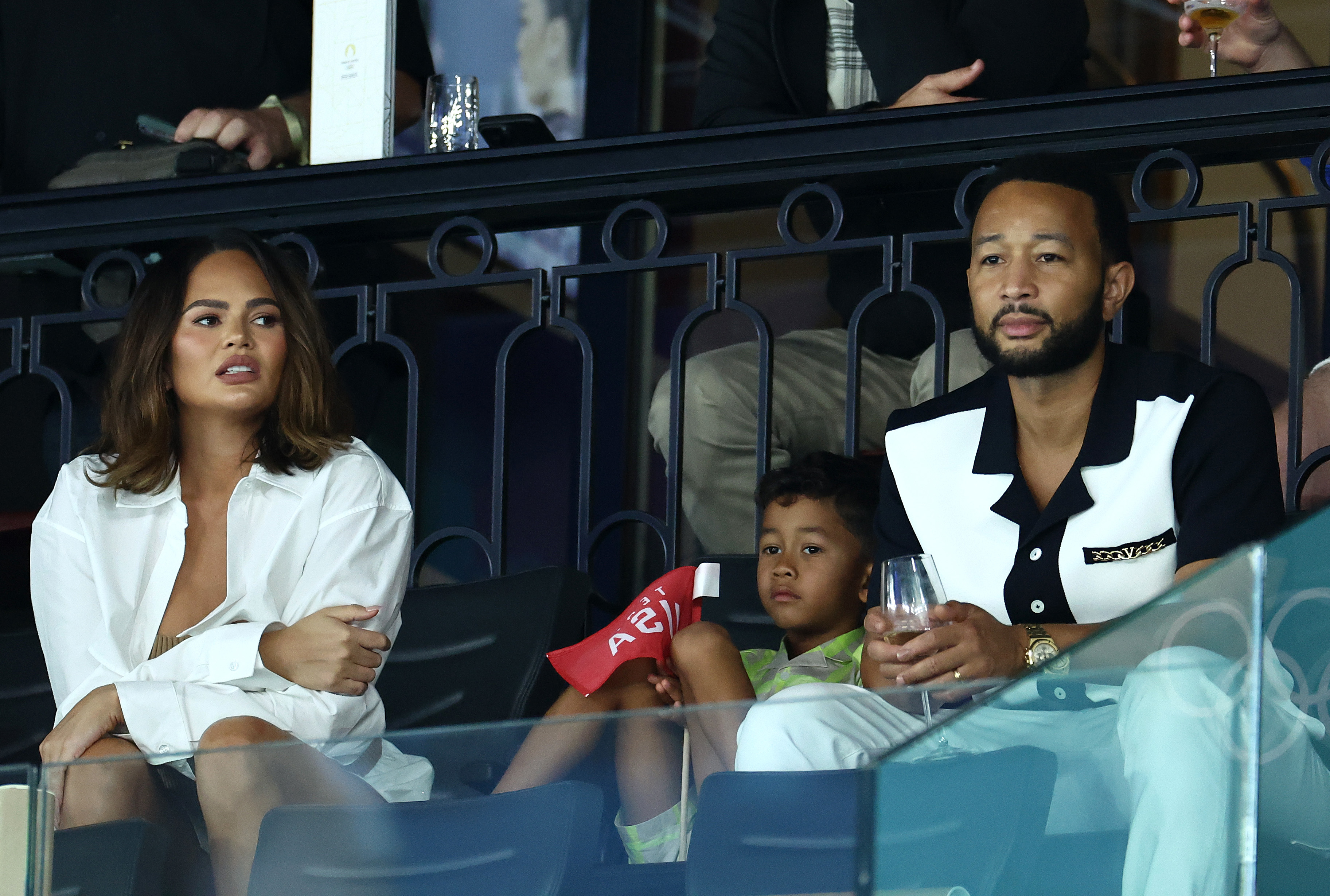 Chrissy Teigen and John Legend are seated with their child at an event, both dressed in stylish outfits, with John holding a drink and their child holding a flag