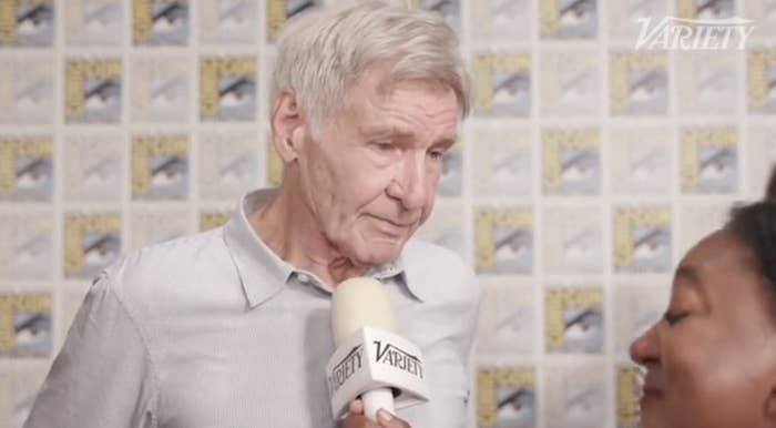 Harrison Ford being interviewed by a Variety reporter at an event with a background featuring multiple poster images. Ford is wearing a light-colored shirt