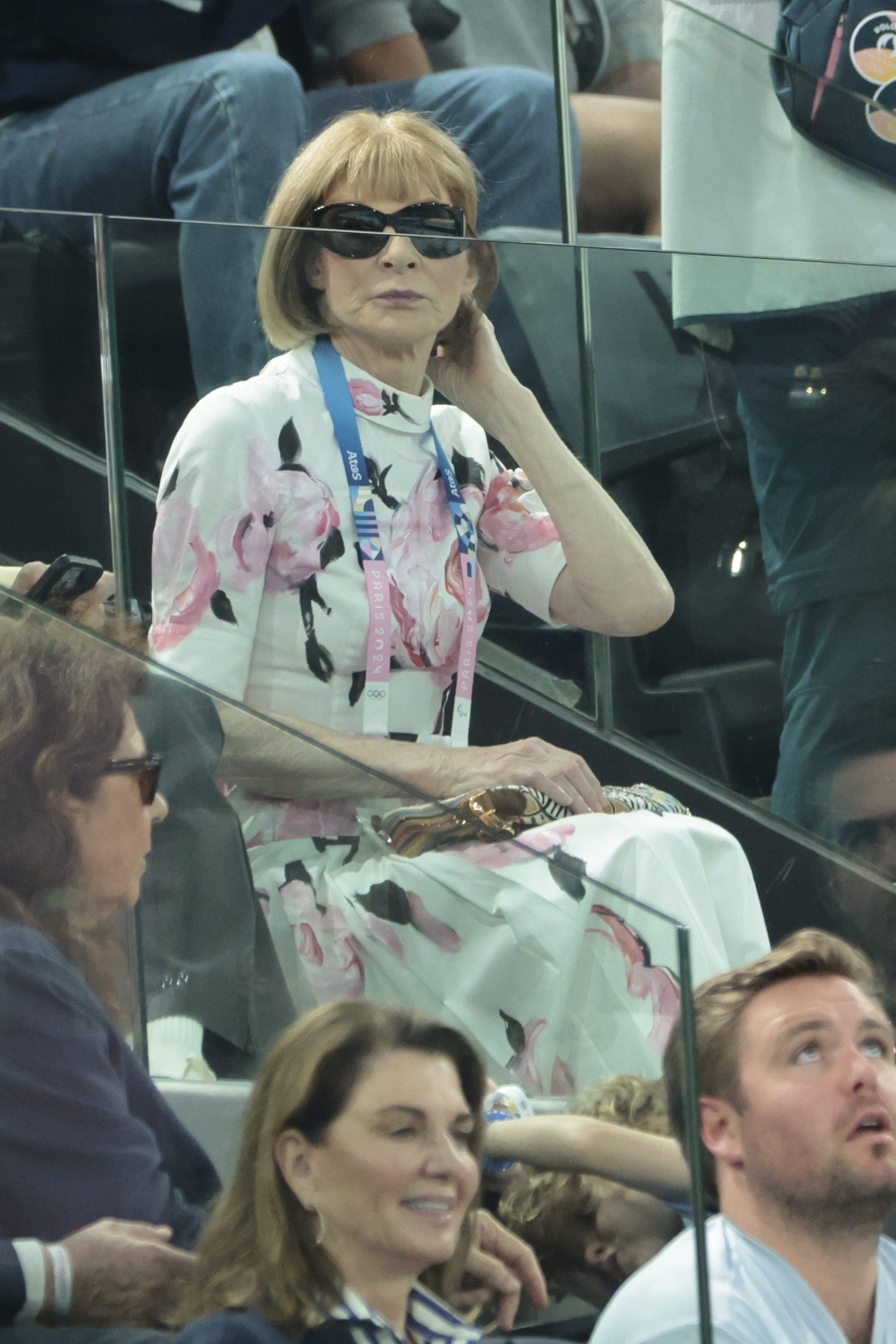 Anna Wintour sits in a stadium box wearing a floral dress and sunglasses, observing the event. Others are seated around her