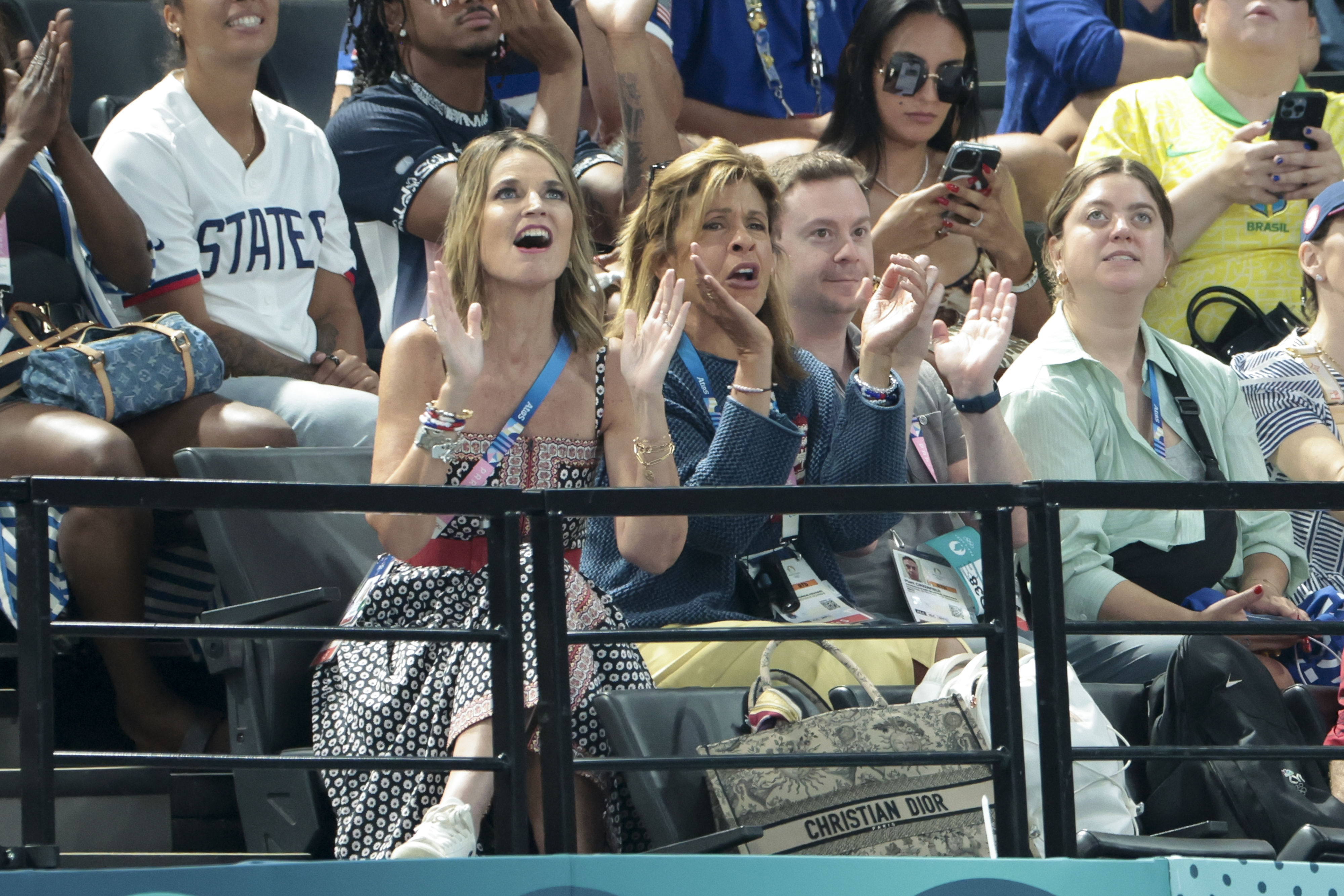 Savannah Guthrie and Hoda Kotb enthusiastically cheer in the stands at an event, wearing casual yet stylish attire while spectators around them capture moments on their phones