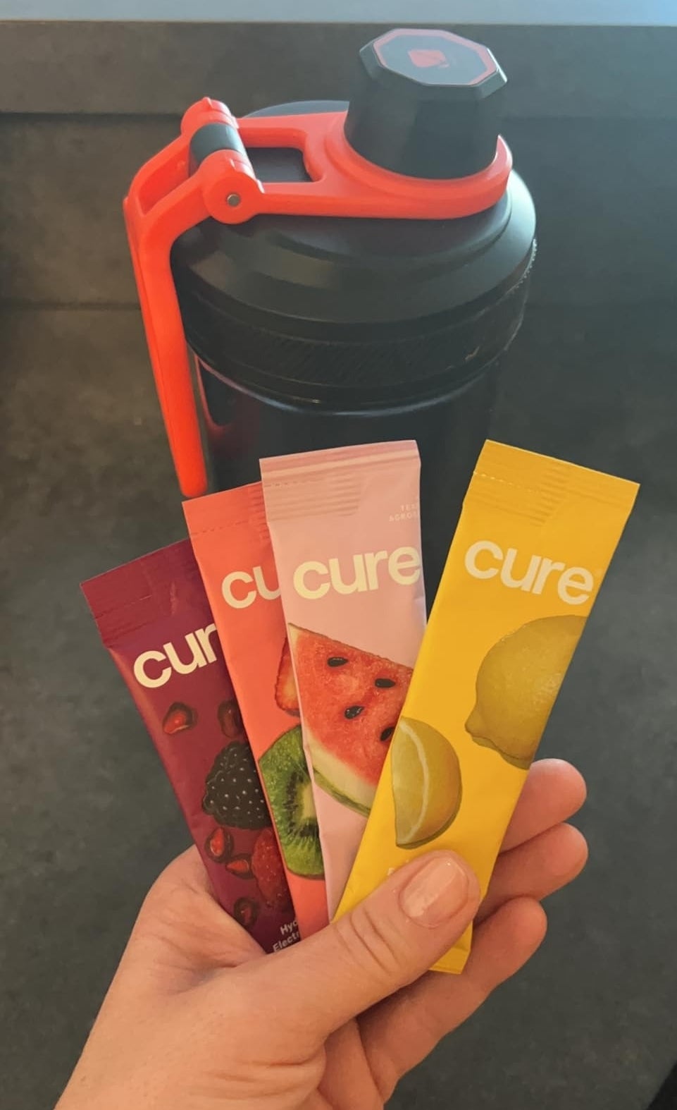 A hand holds four Cure hydration sachets in different flavors (berry, watermelon, and lemon) in front of a sports water bottle