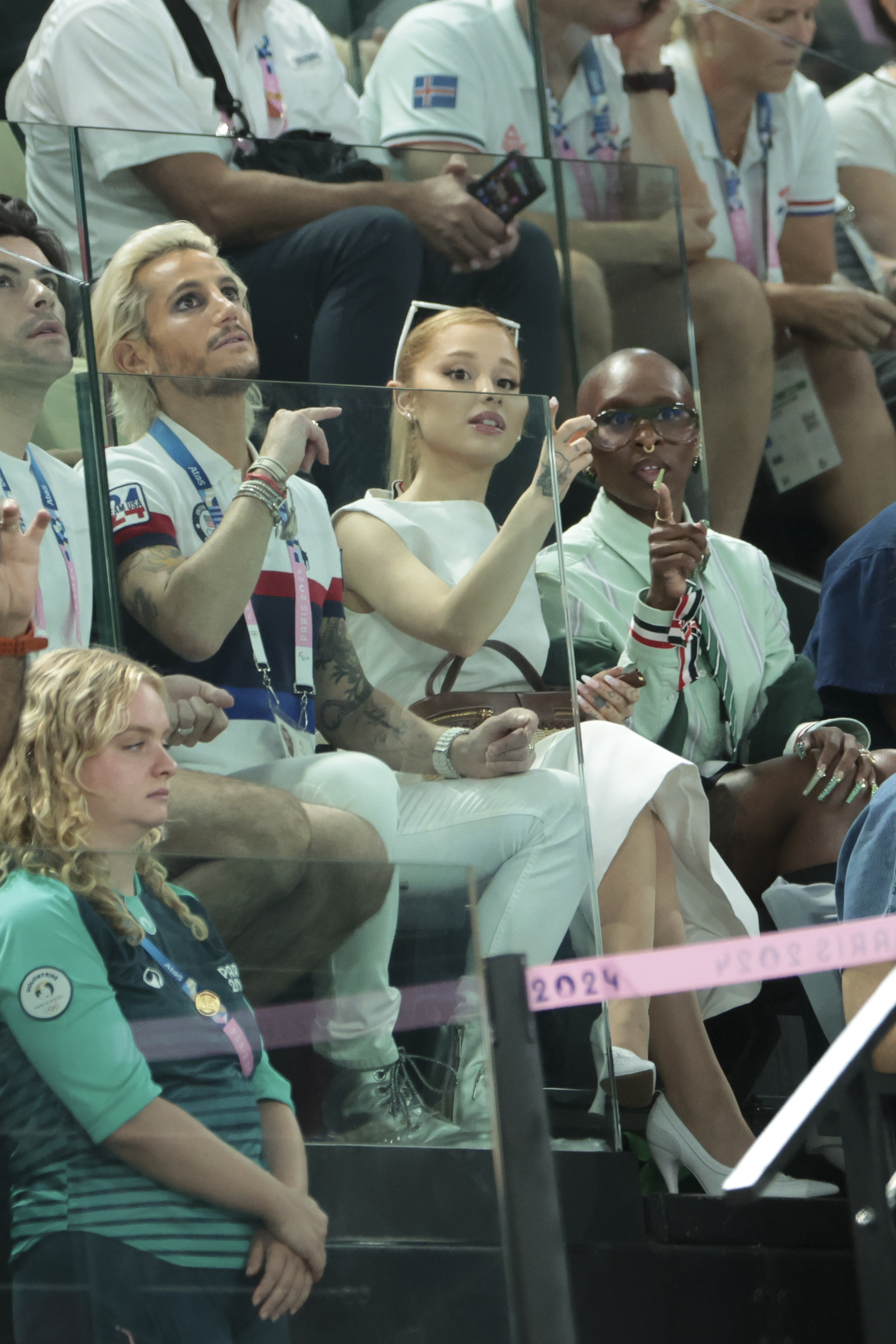 Ariana Grande, Frankie Grande, and Jonathan Bailey seated in a sports arena audience, with Ariana and Jonathan pointing towards something in front