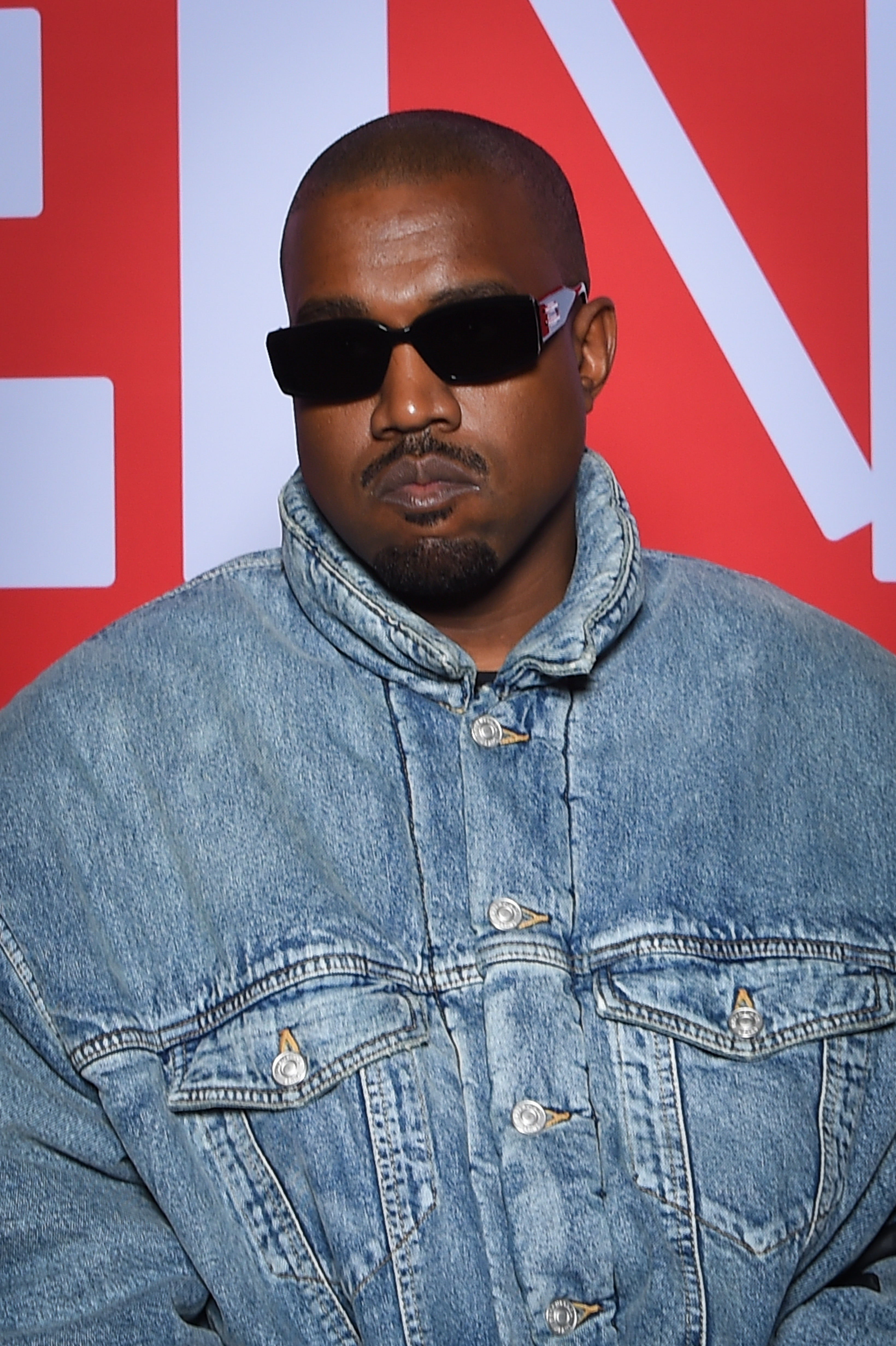 Kanye West in a denim jacket and dark sunglasses, standing against a backdrop with large text