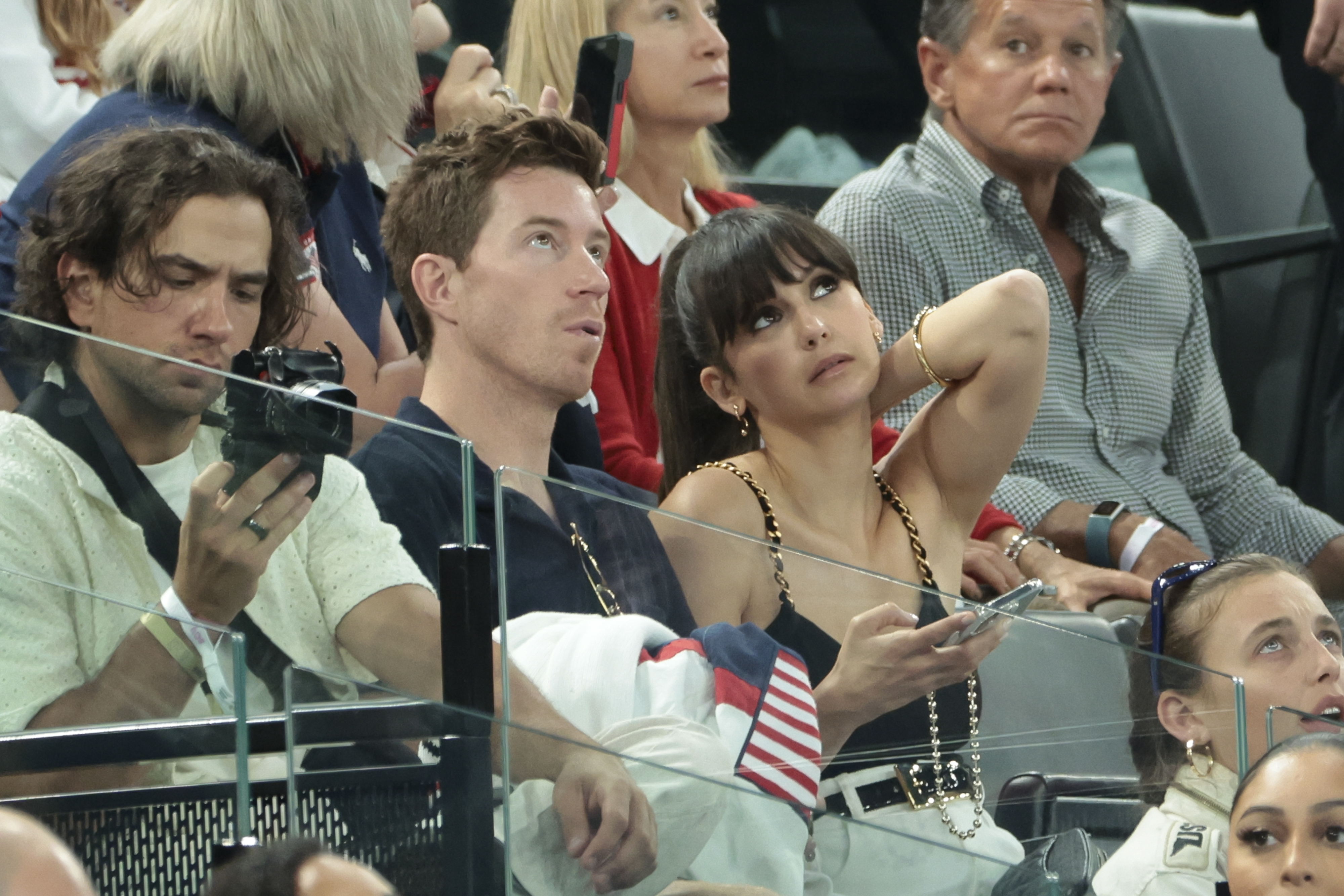 From left to right: Daren Kagasoff, Shaun White, and Nina Dobrev sitting together in the stands, with Dobrev holding her phone and looking up