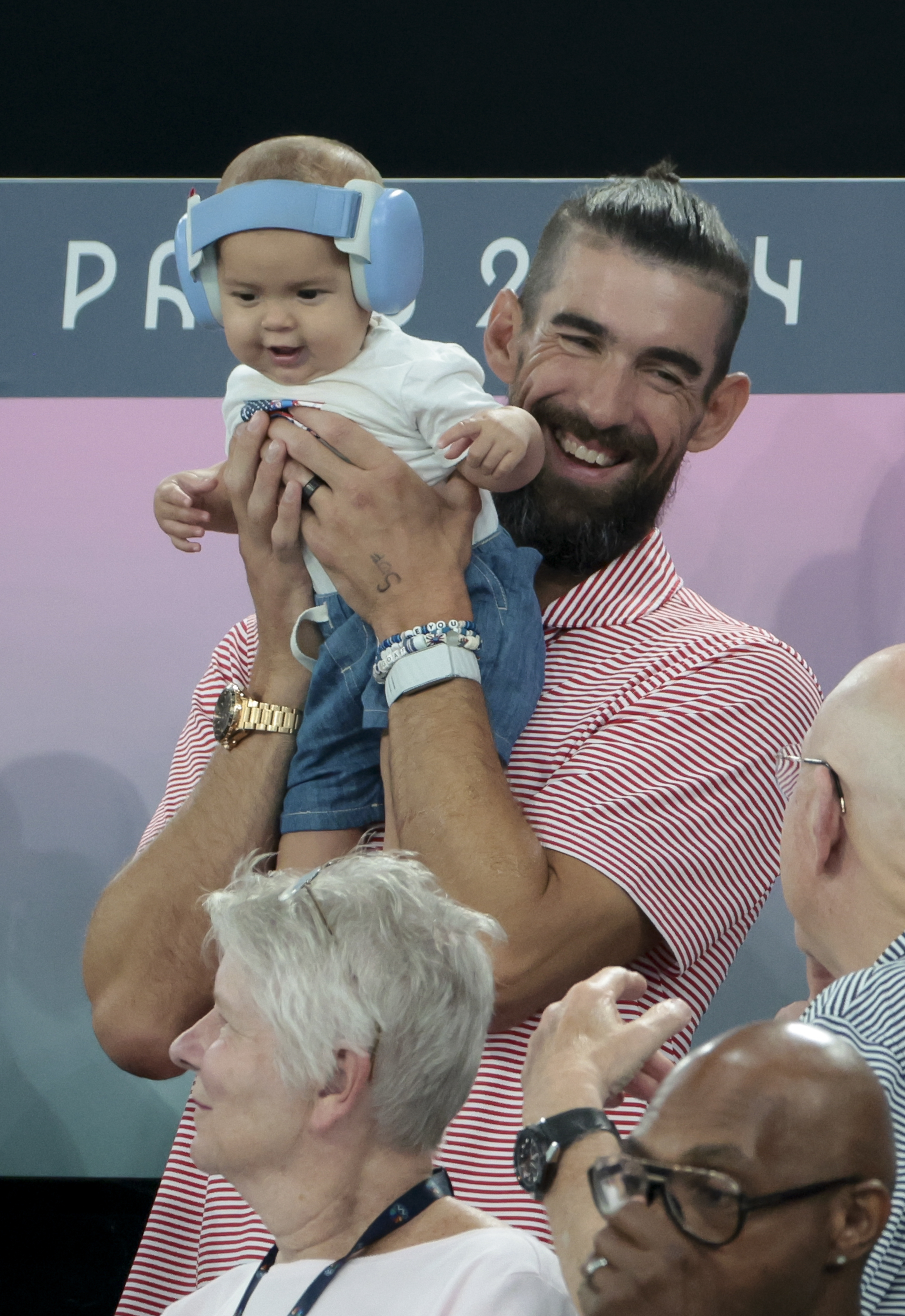 Michael Phelps, smiling in a striped shirt, holds a baby wearing earmuffs. People are chatting in the foreground