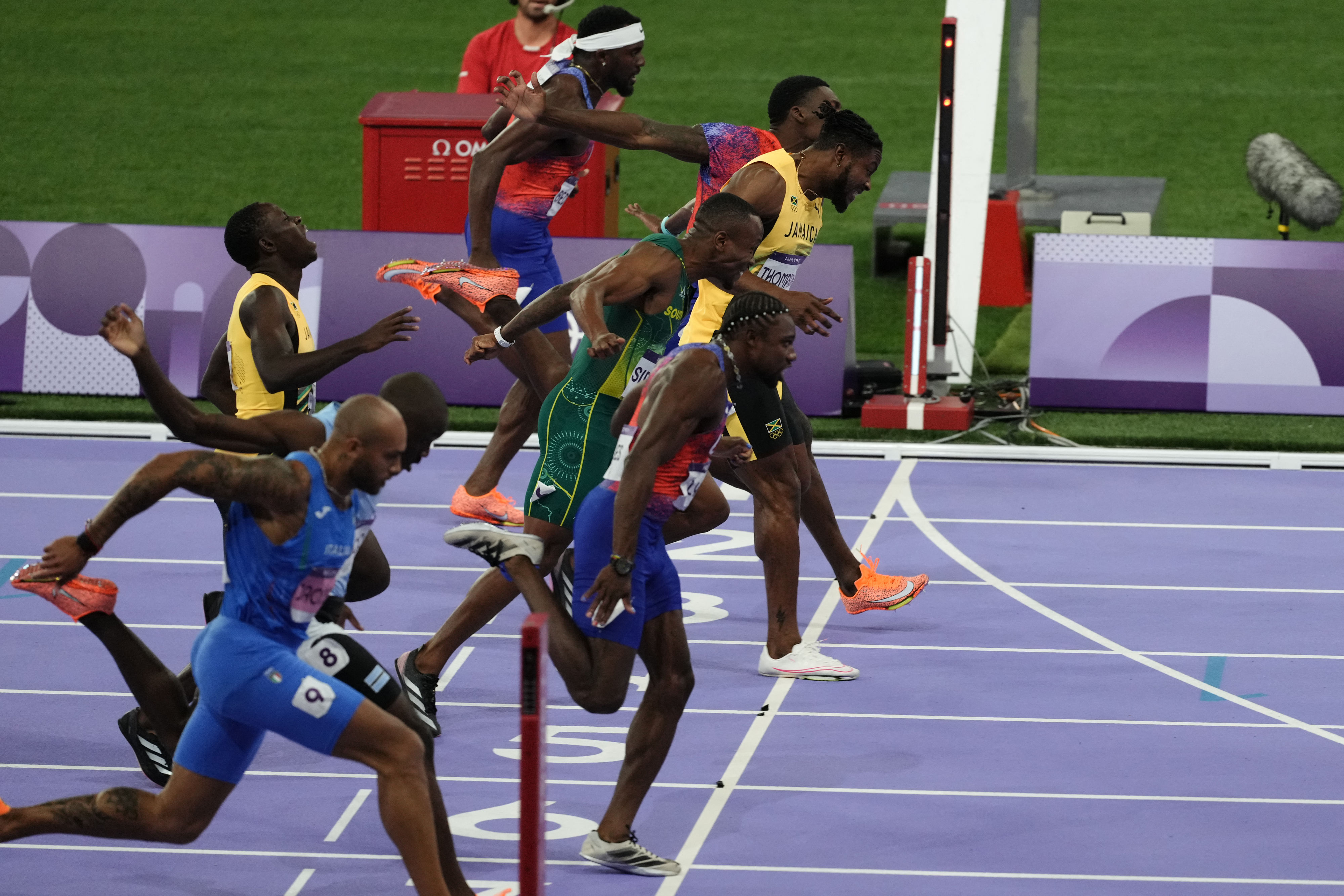 Sprinters cross the finish line during a track race at a stadium. The athletes are wearing race attire and looking focused