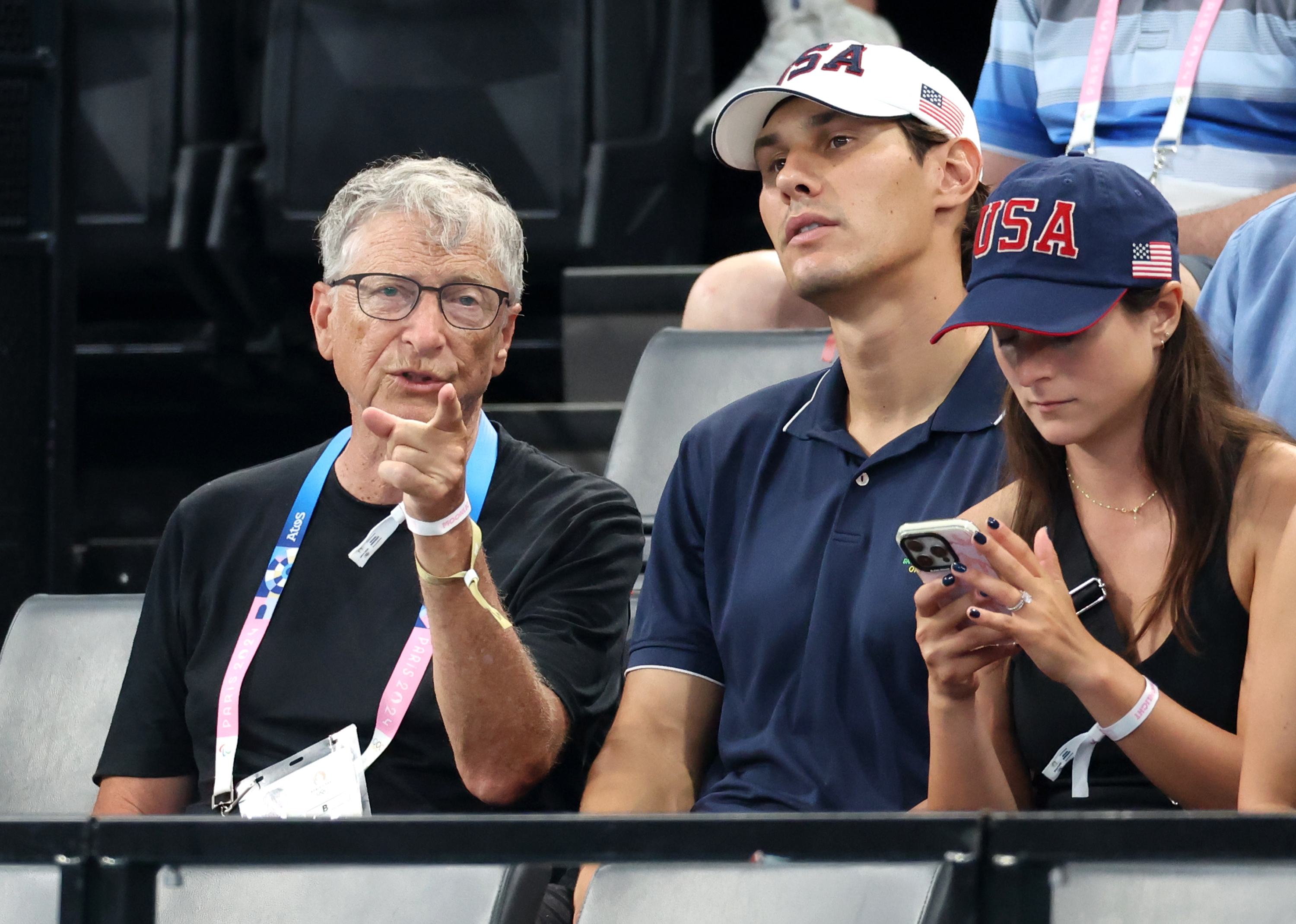 Bill Gates, a man in a USA hat and polo shirt, and a woman in a USA hat at a sports event. Bill Gates is pointing, and the woman is using a smartphone