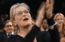 Meryl Streep aplaudiendo en lo que parece ser un evento formal. Personas al fondo también aplauden