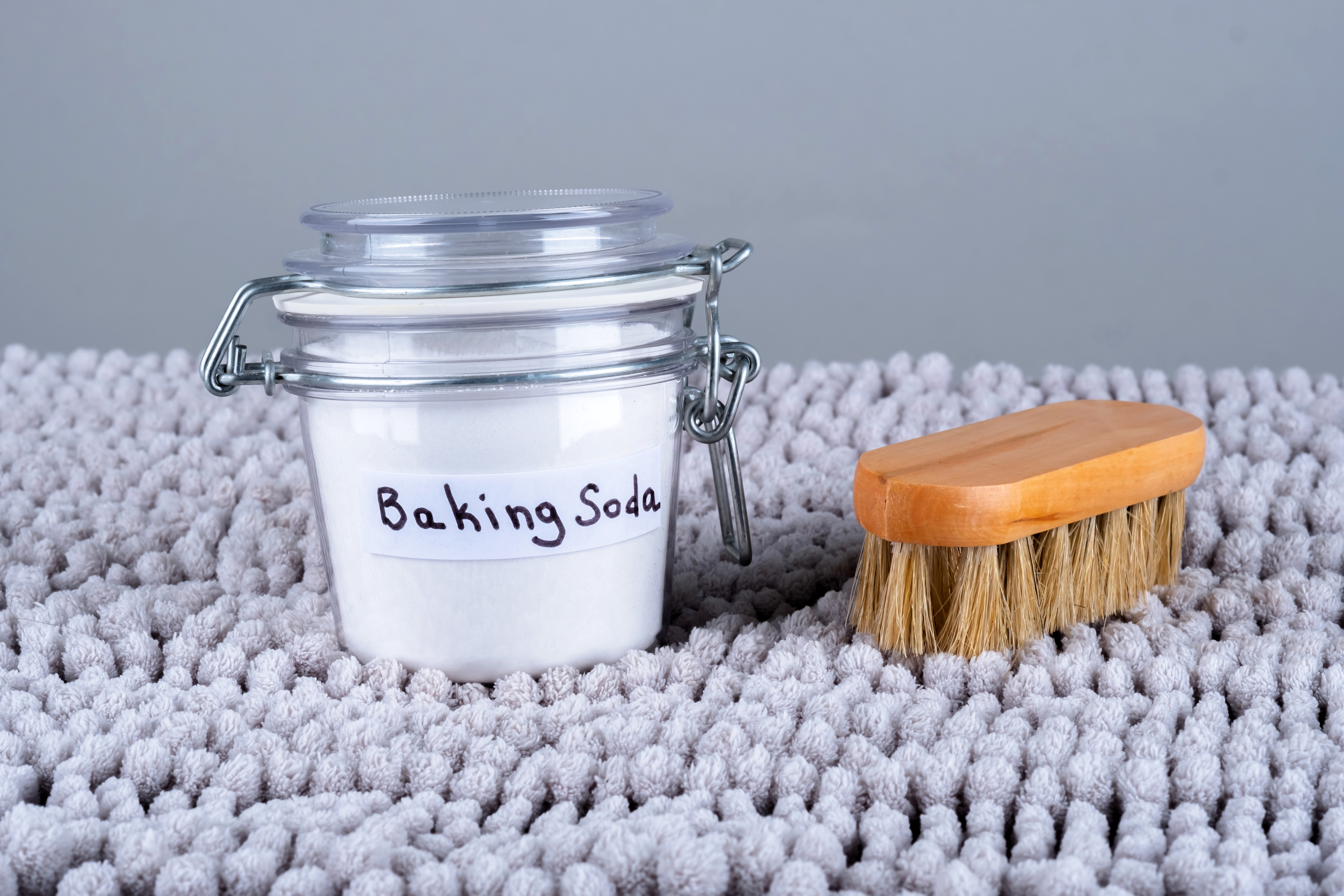 A jar labeled "Baking Soda" and a brush with a wooden handle are placed on a textured surface