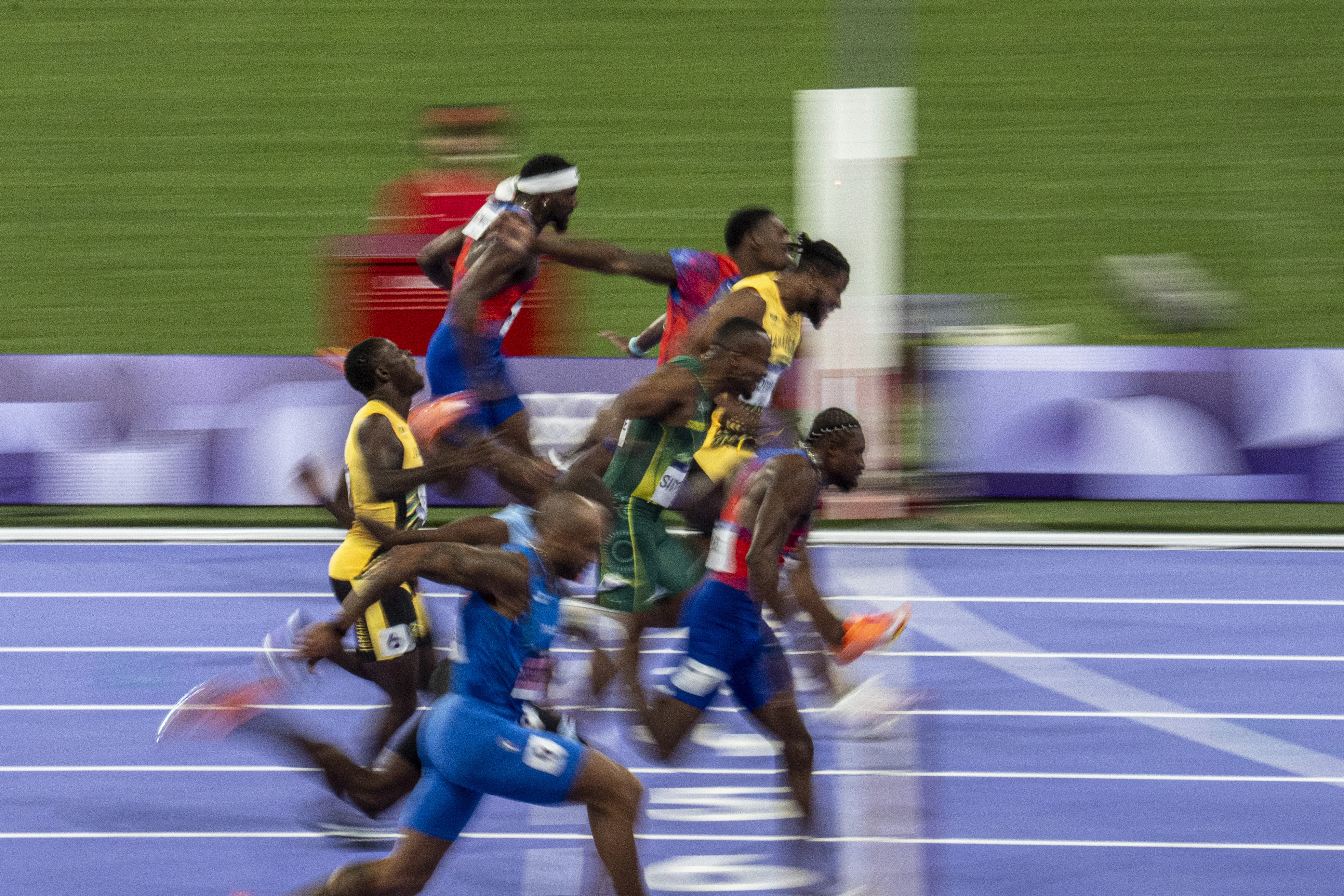 Track athletes sprinting towards the finish line, captured mid-motion. They are all leaning forward intensely, striving to cross the line first in a close race